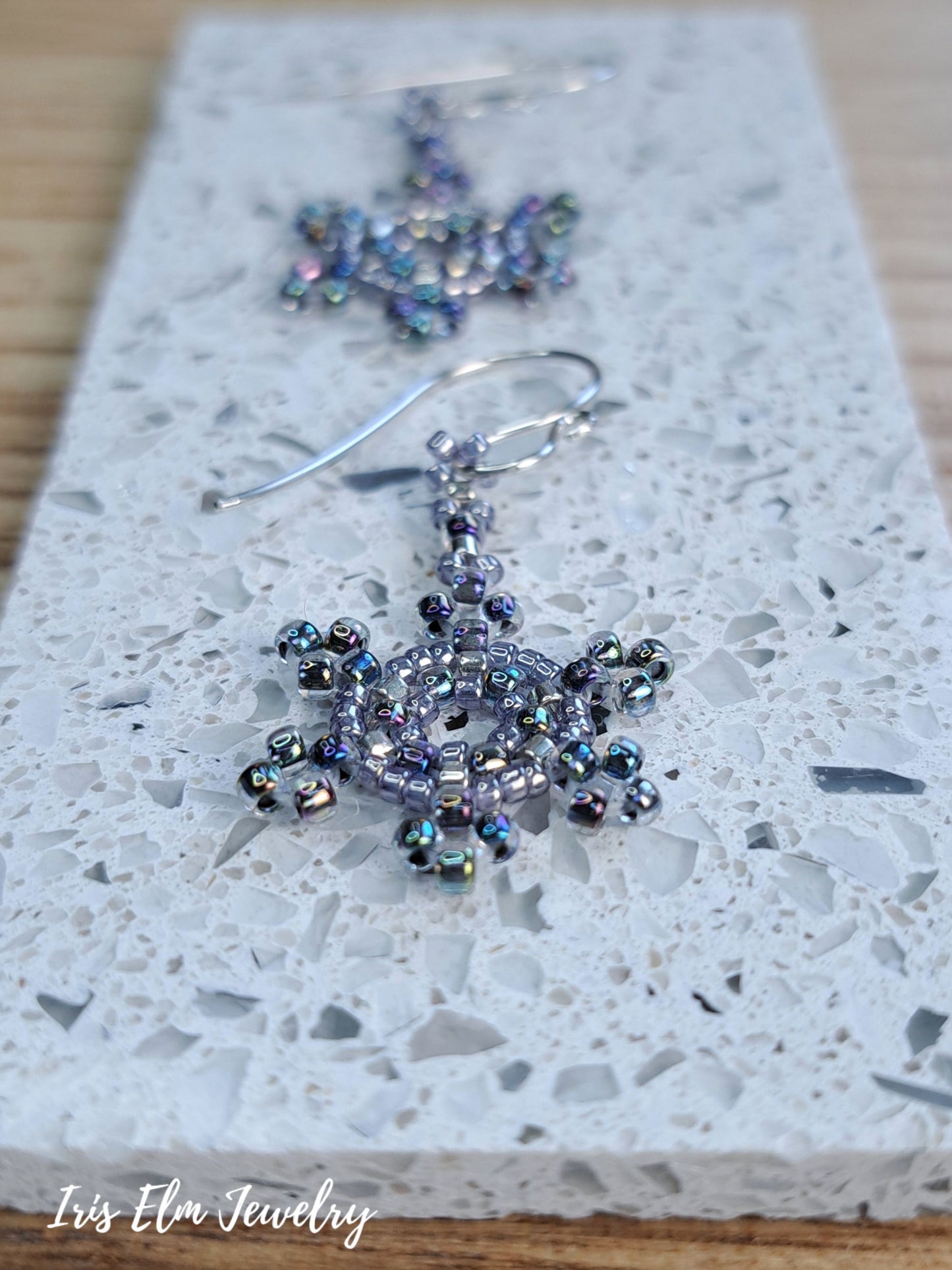 Snowflake-shaped beaded drop earrings with iridescent blue and purple beads on silver fishhook wires, displayed on a white speckled stone tile with the brand name 'Iris Elm Jewelry' in the bottom-left corner.