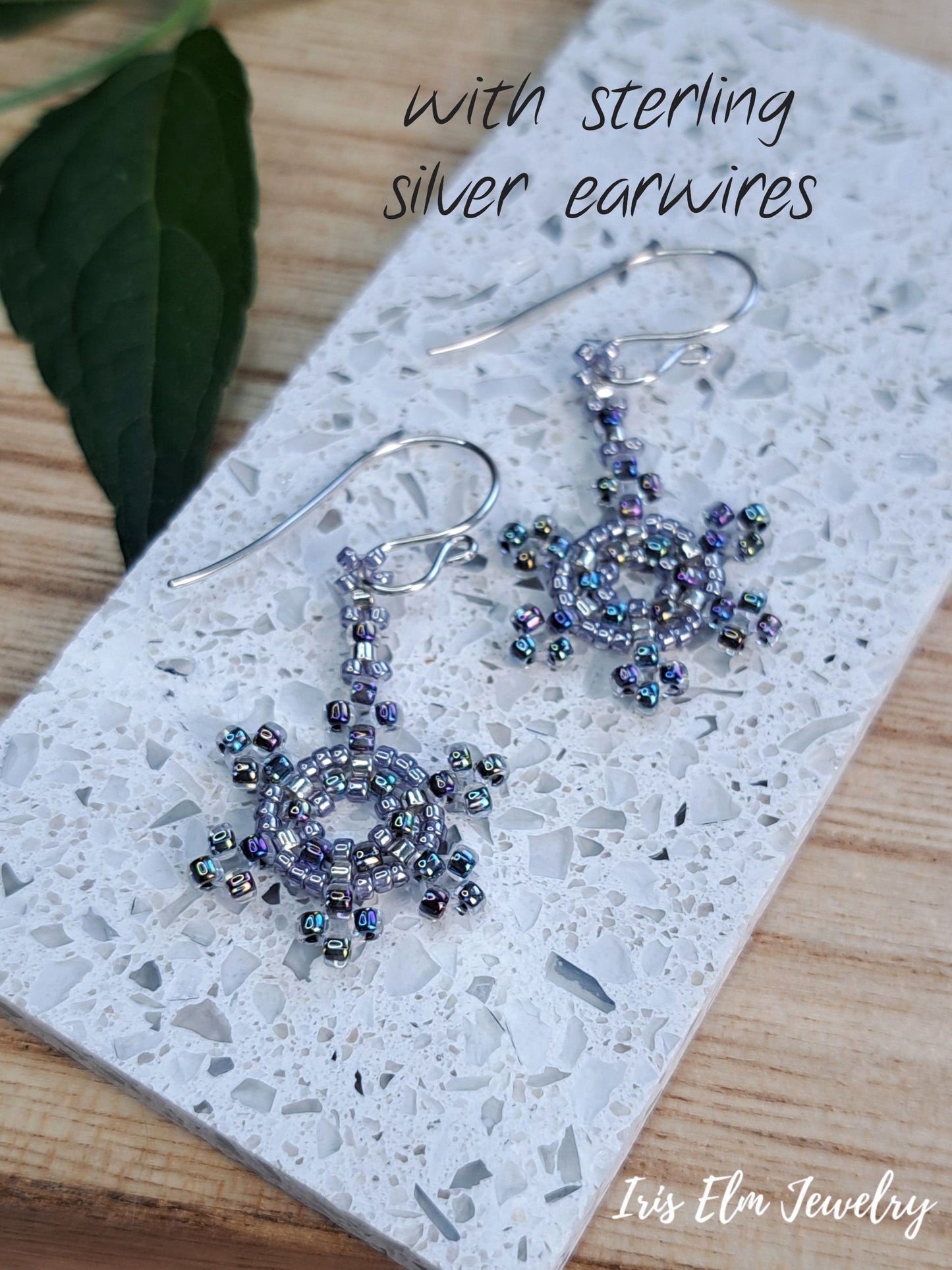 A pair of handmade beaded dangle earrings with silver fishhook earwires, displayed on a white speckled stone tile over a wooden surface. The earrings feature intricate purple and iridescent blue beads.