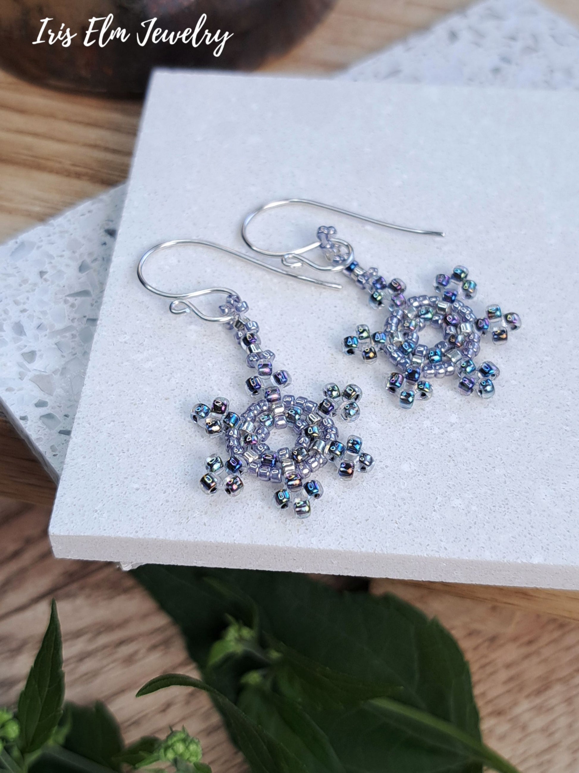 A pair of handmade beaded dangle earrings by Iris Elm Jewelry, featuring silver fishhook wires and circular snowflake-like clusters of iridescent lavender and blue beads, displayed on a white textured block.