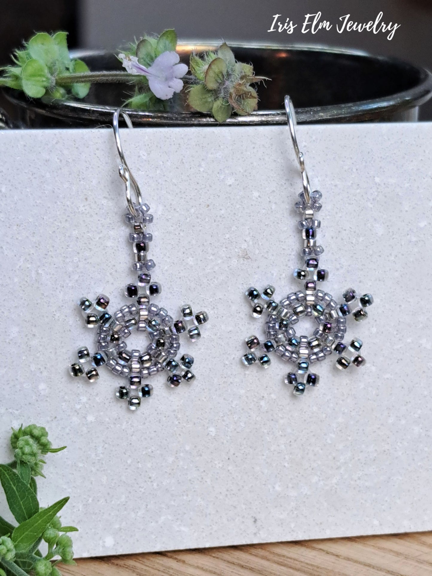 A pair of handmade beaded dangle earrings with a snowflake-like circular design, displayed on a textured white card. The earrings are made with silver-toned metal hooks and beads that shimmer in silver, gray, and iridescent blue-green.