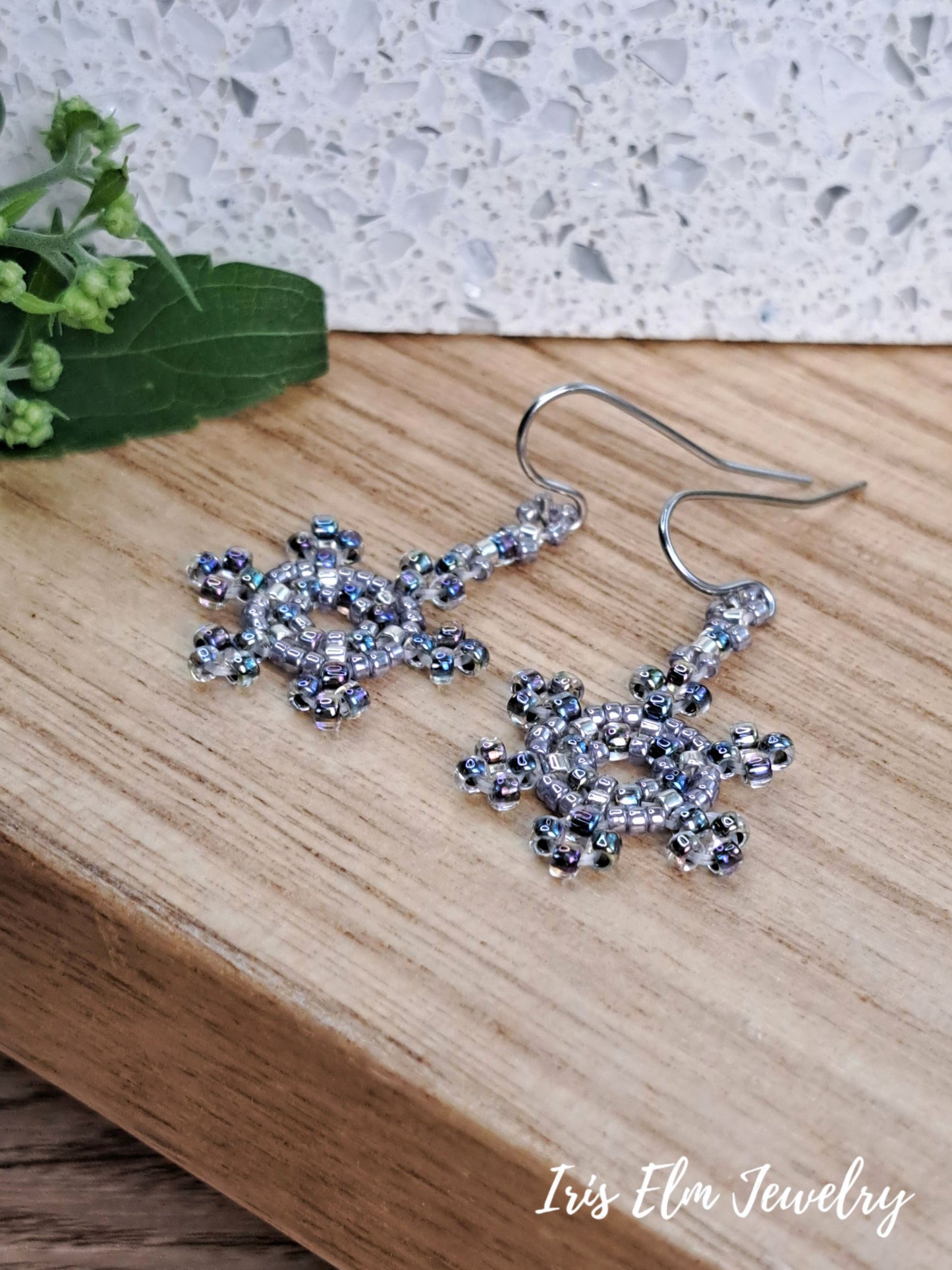 A pair of silver-toned dangle earrings with intricate beaded snowflake-like clusters of clear and iridescent rhinestones, resting on a light wood surface with a white speckled background and green leaves in the corner. The brand name 'Iris Elm Jewelry' is visible in the bottom right corner.