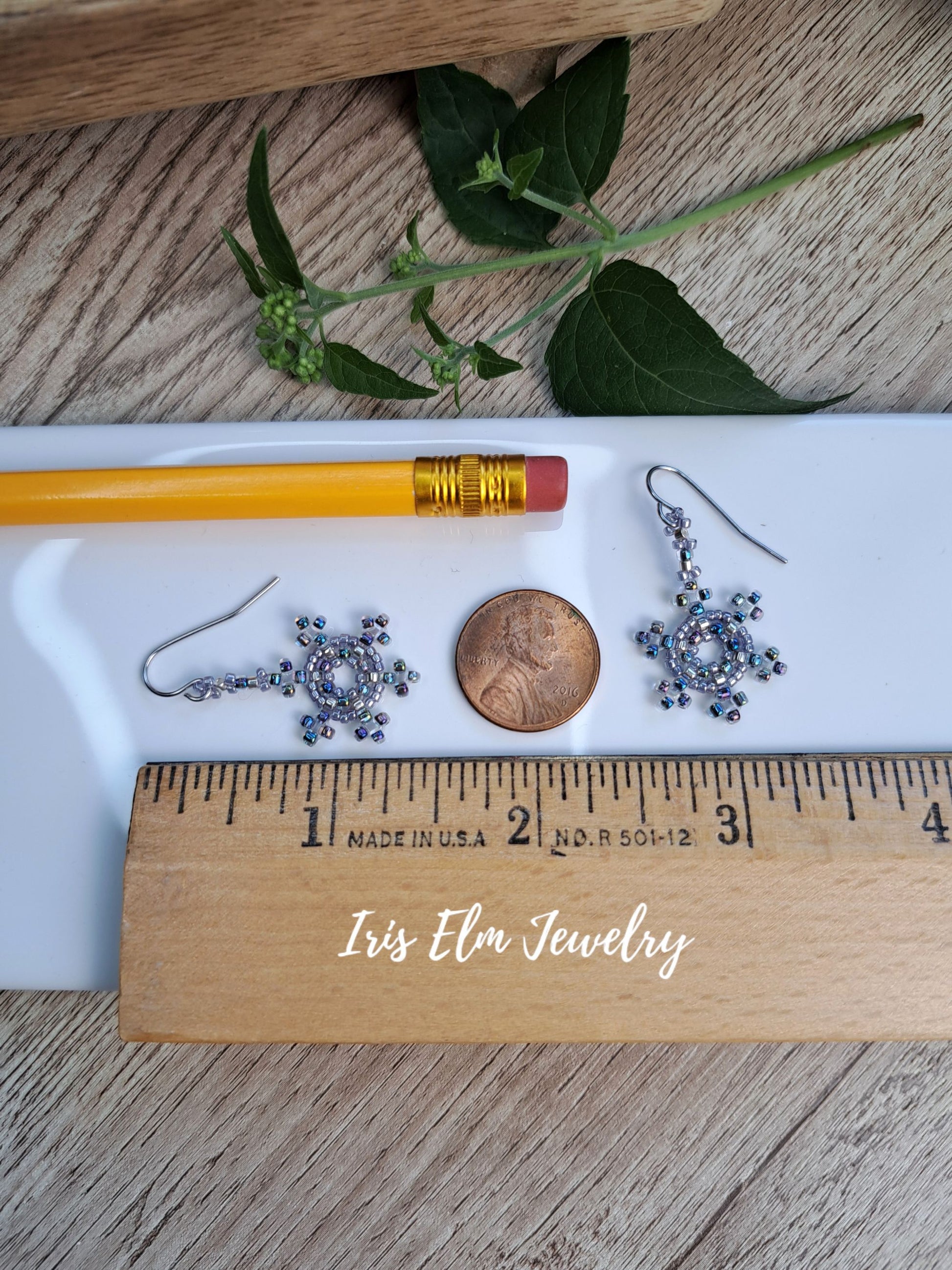 A pair of handmade beaded dangle earrings with silver hooks and blue-gray beads, displayed on a white tray with a pencil, penny, wooden ruler, and green leaves on a wood surface. The text 'Iris Elm Jewelry' is written on the wooden ruler.
