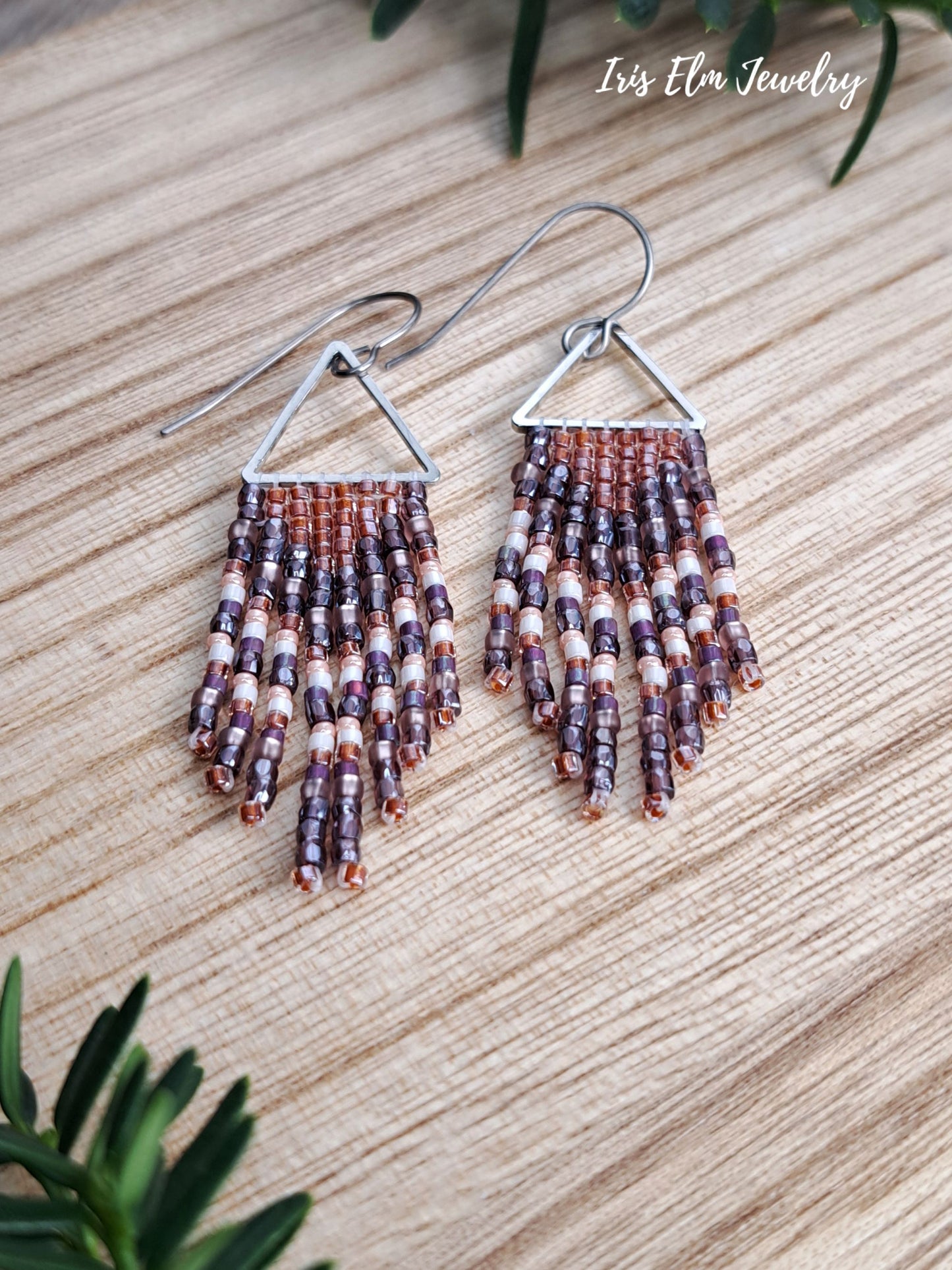 A pair of handmade beaded dangle earrings with silver triangular tops and silver fishhook wires, featuring beads in brown, purple, and cream. The earrings are displayed on a light wood grain surface with green leaves in the corners and the text 'Iris Elm Jewelry' in the top right.