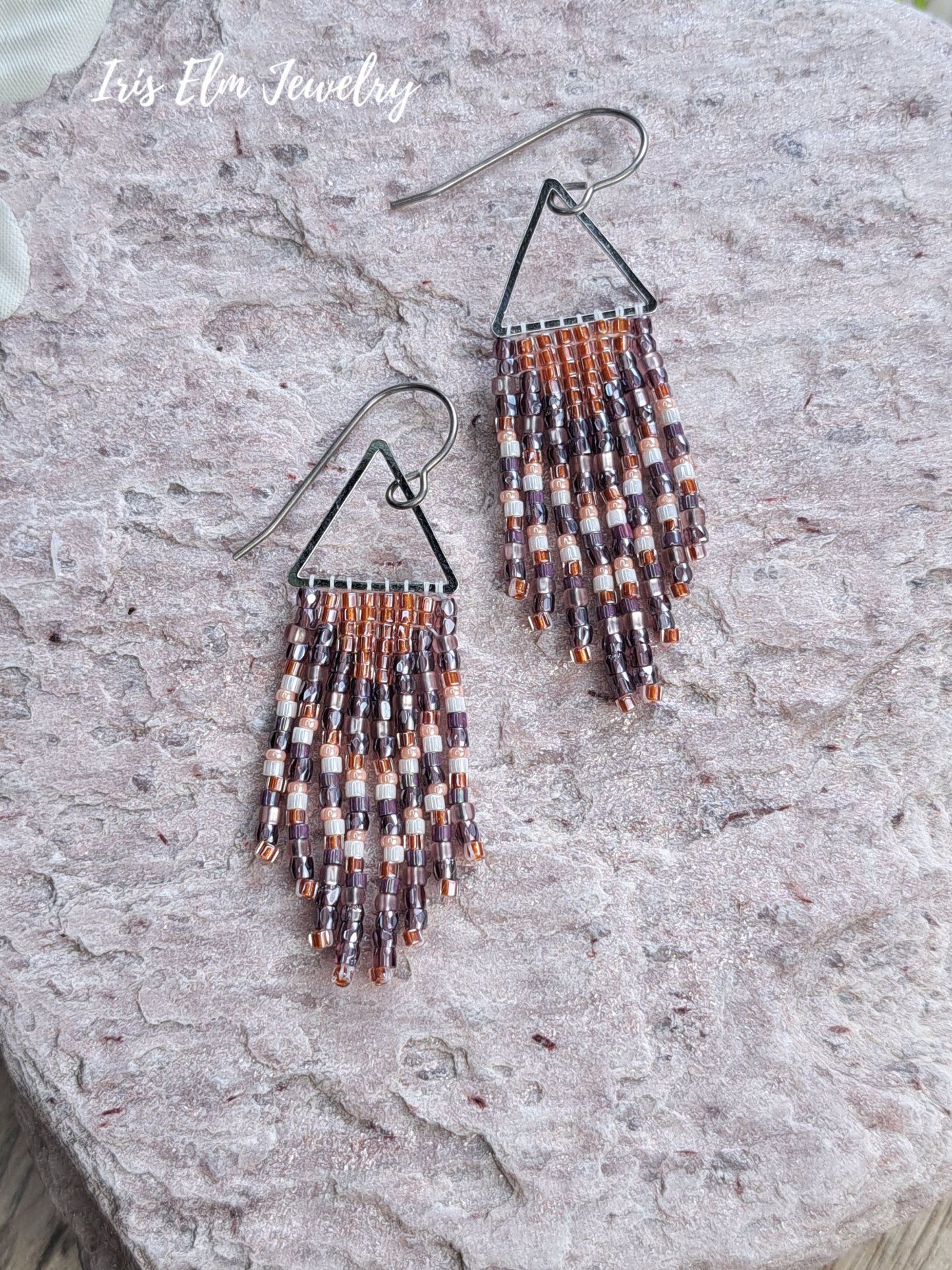 handmade small seed bead handwoven purple and copper orange seed bead fringe earrings on rocky surface with Iris Elm Jewelry branding.