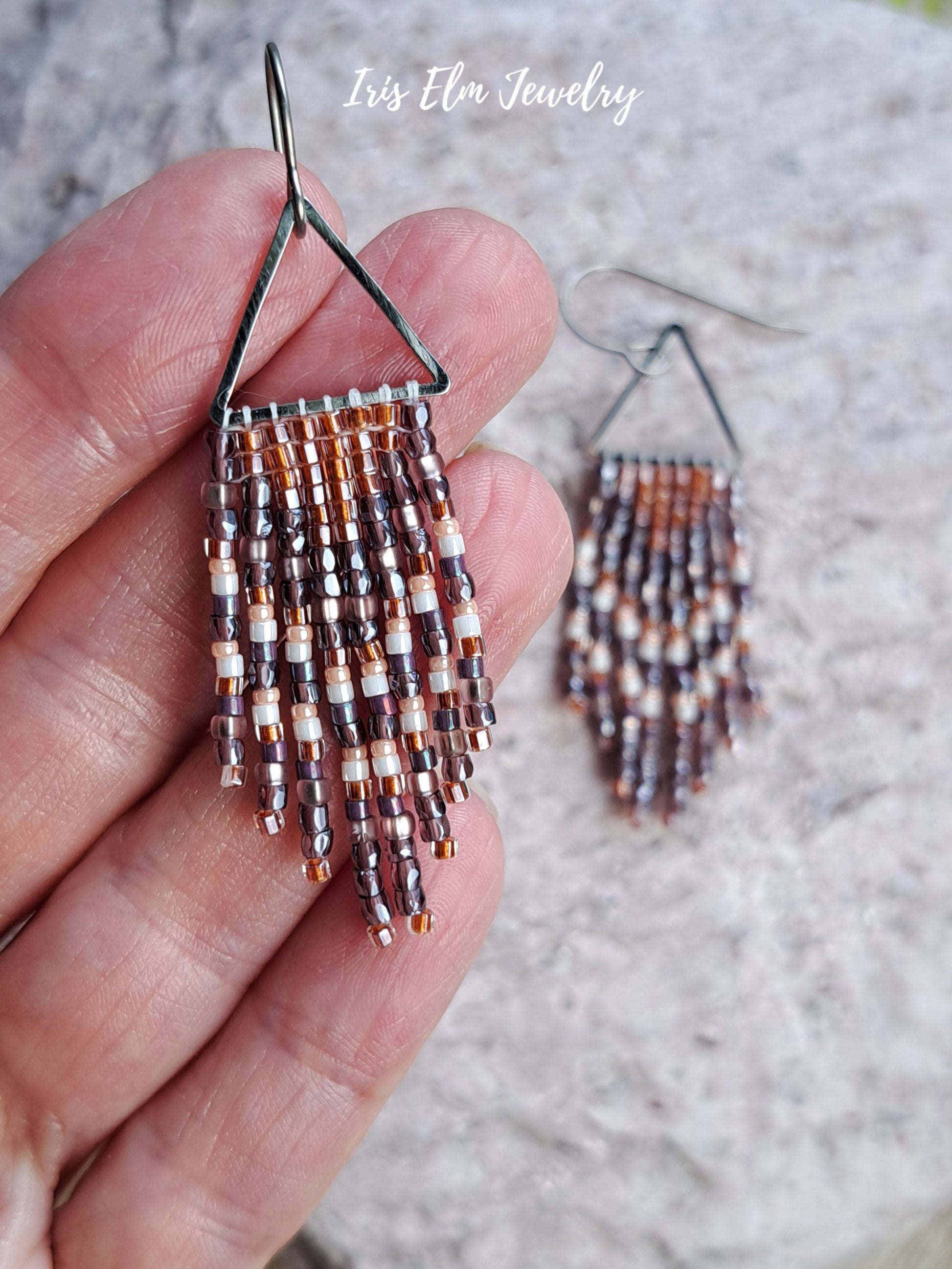 handmade small seed bead handwoven purple and copper orange seed bead triangle fringe earrings held by hand with Iris Elm Jewelry branding.