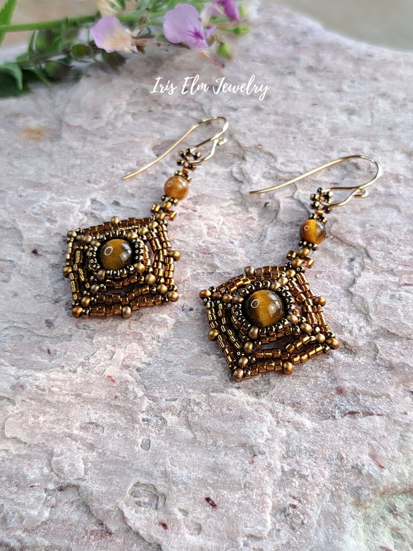 A pair of handmade beaded dangle earrings with square bronze and gold geometric frames, featuring a central round brown tiger eye stone and a small tiger eye bead above, displayed on a textured light gray stone surface with a watermark reading 'Iris Elm Jewelry'.