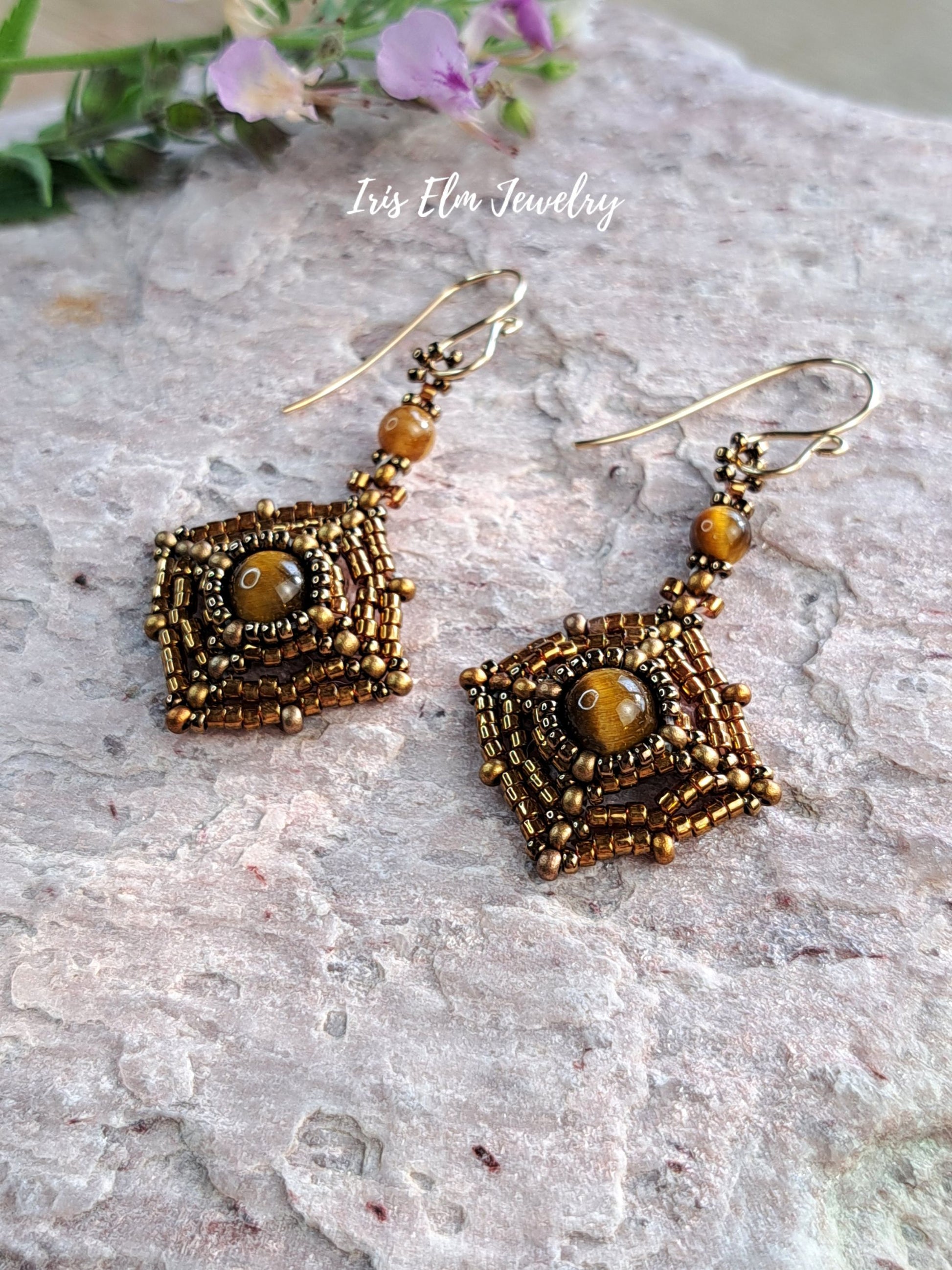 A pair of handmade beaded dangle earrings with square bronze and gold geometric frames, featuring a central round brown tiger eye stone and a small tiger eye bead above, displayed on a textured light gray stone surface with a watermark reading 'Iris Elm Jewelry'.