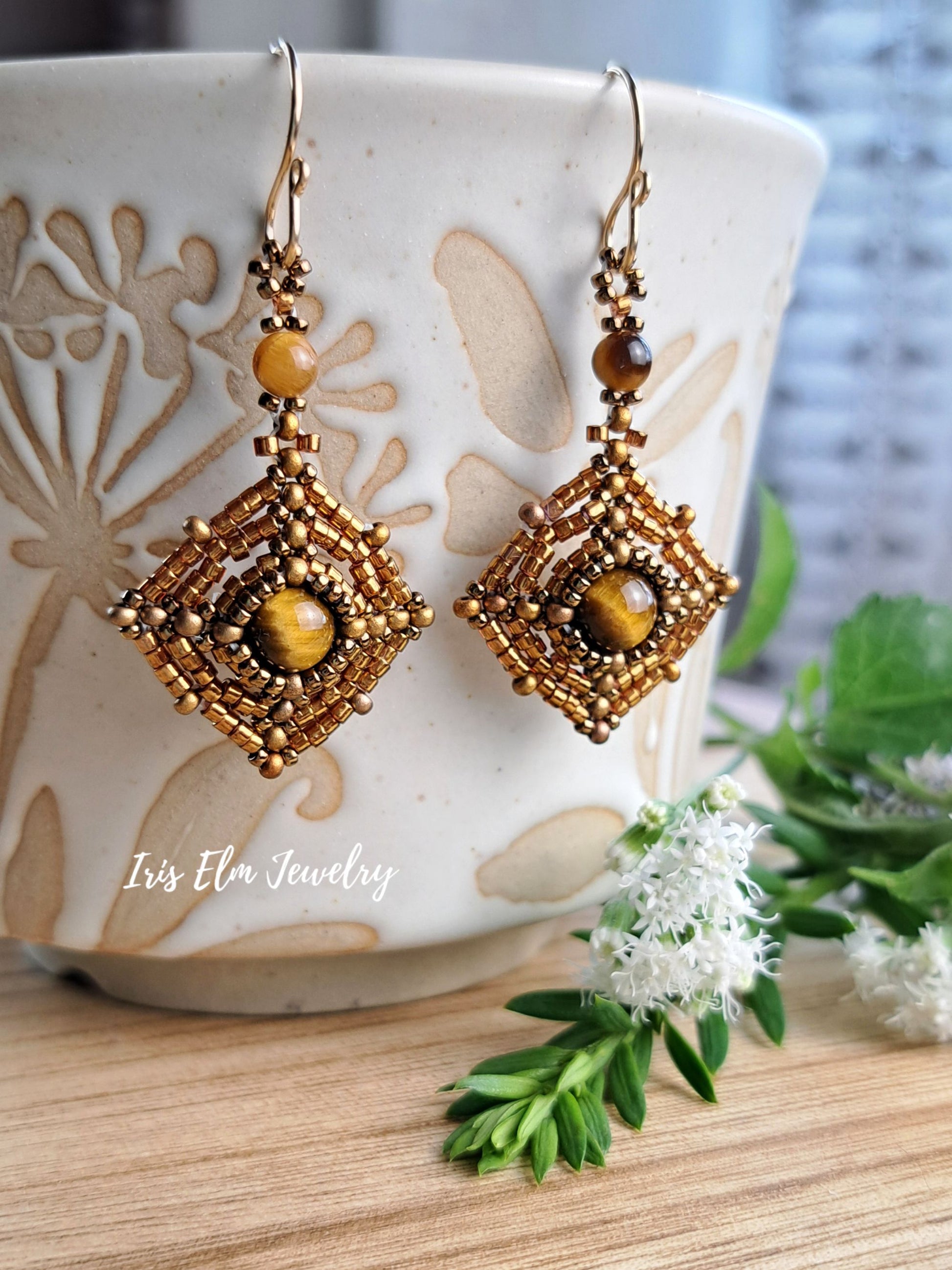 A pair of handmade bohemian-style beaded earrings with gold-toned metal, brown beads, and tiger's eye stones, hanging from a ceramic cup on a wooden surface with green leaves and white flowers.