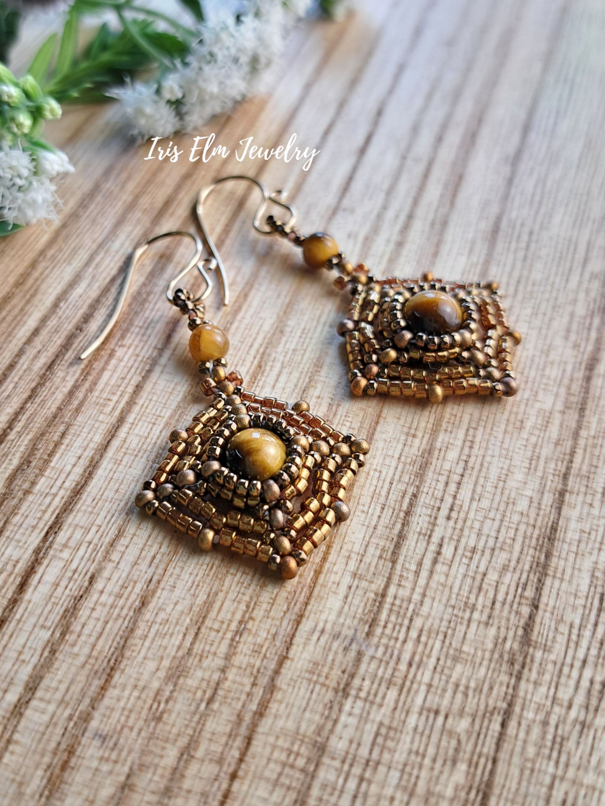 A pair of handmade beaded dangle earrings with square geometric patterns, featuring brown and gold beads, tiger's eye center stones, and fishhook wires. They are displayed on a light wood grain surface with a watermark reading 'Iris Elm Jewelry'.
