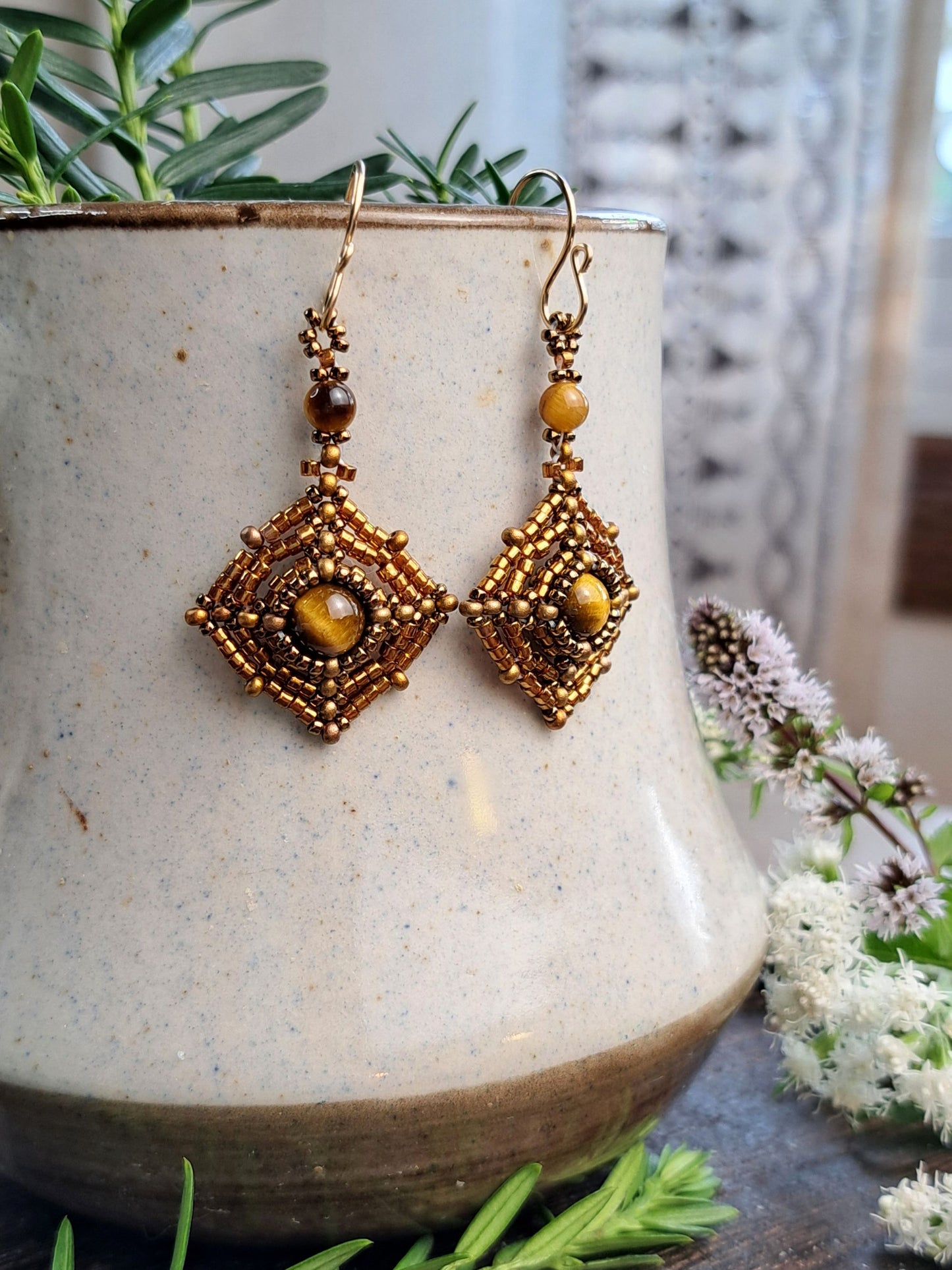 A pair of bohemian-style gold and brown beaded dangle earrings with tiger's eye stones, hanging on a speckled ceramic planter with green foliage and white flowers.
