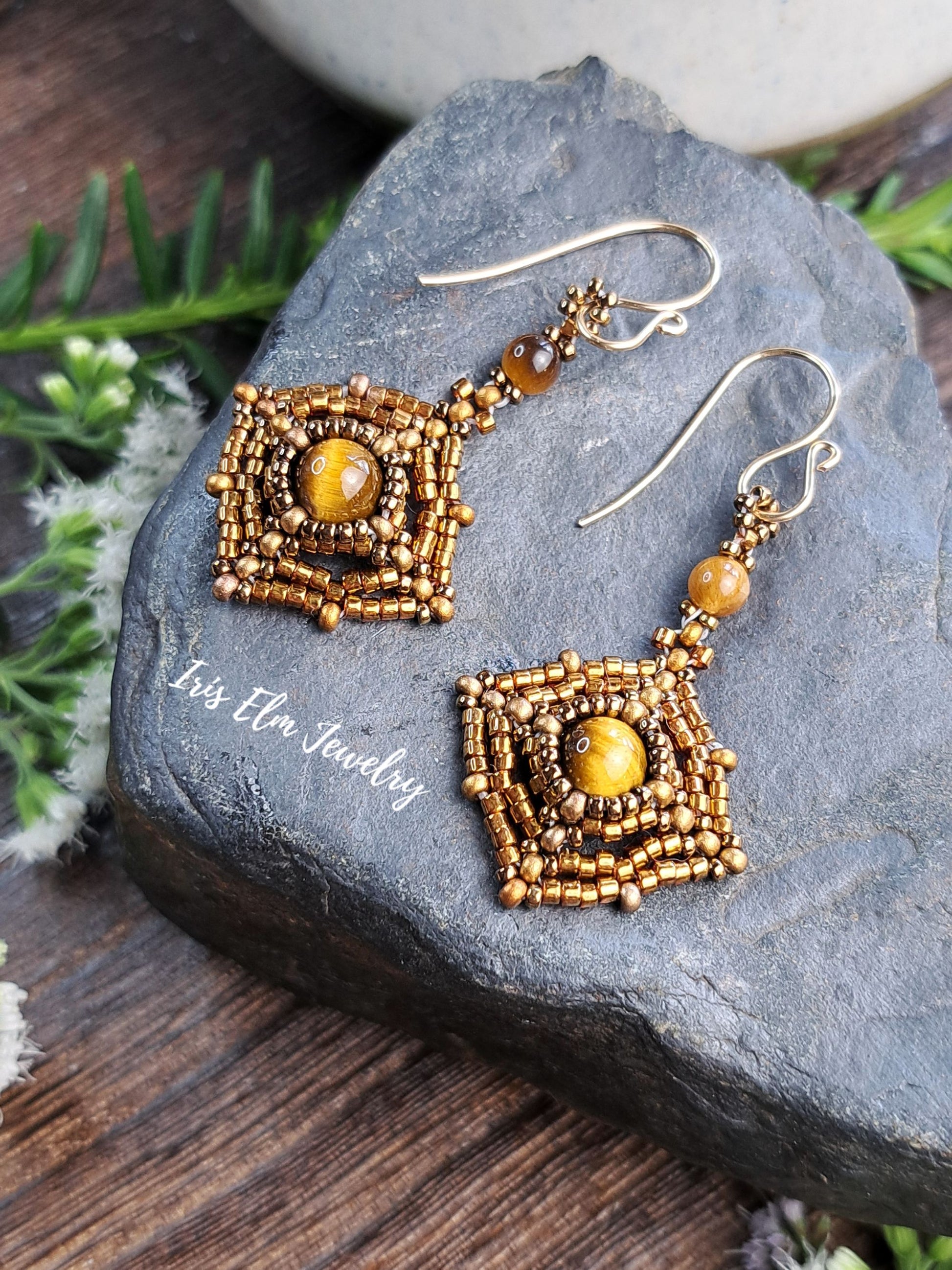 A pair of handmade beaded dangle earrings with square gold-toned beadwork and amber-yellow center beads, displayed on a dark stone surface with a wooden background and small greenery.