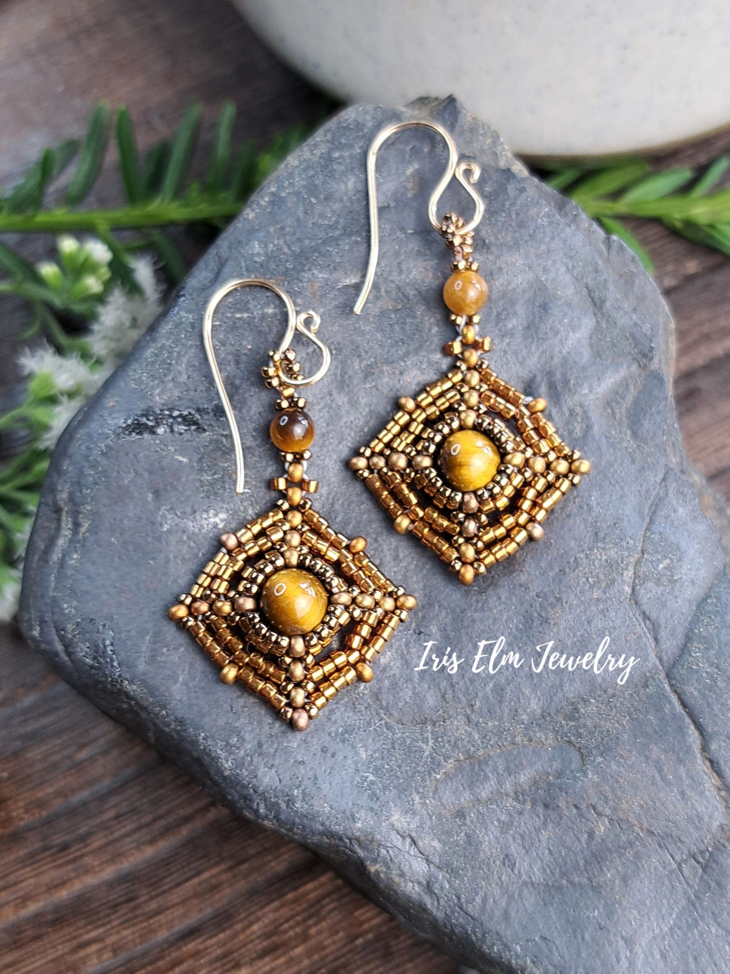 A pair of handmade beaded dangle earrings with geometric gold and brown patterns, featuring a central tiger eye bead and silver-tone fishhook wires, displayed on a dark slate stone with pine branches.