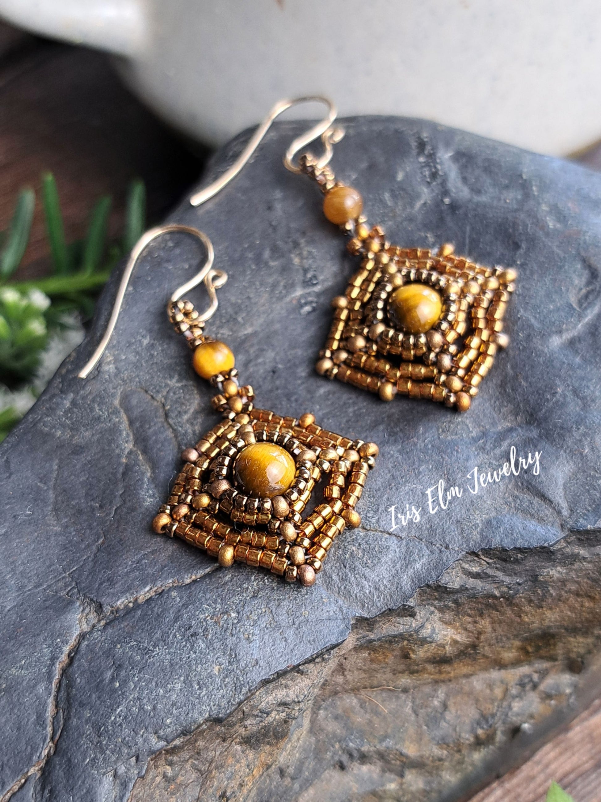 A pair of handmade beaded brown tiger eye dangle earrings with gold-toned fishhook wires and square-shaped drop pendants featuring a central round gold bead, displayed on a dark gray stone surface.