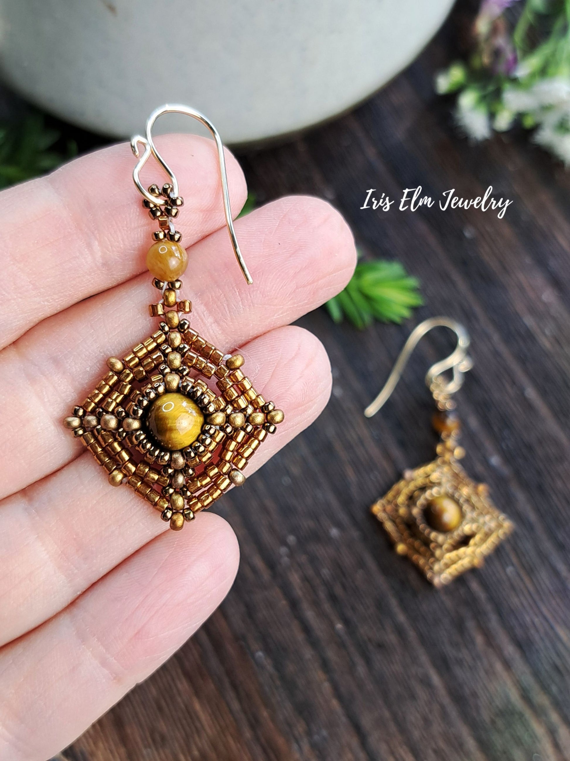 A hand holds a gold and bronze beaded geometric dangle earring with a round tiger's eye bead, next to a matching earring resting on a dark wood surface. The brand name 'Iris Elm Jewelry' is visible in the upper right.