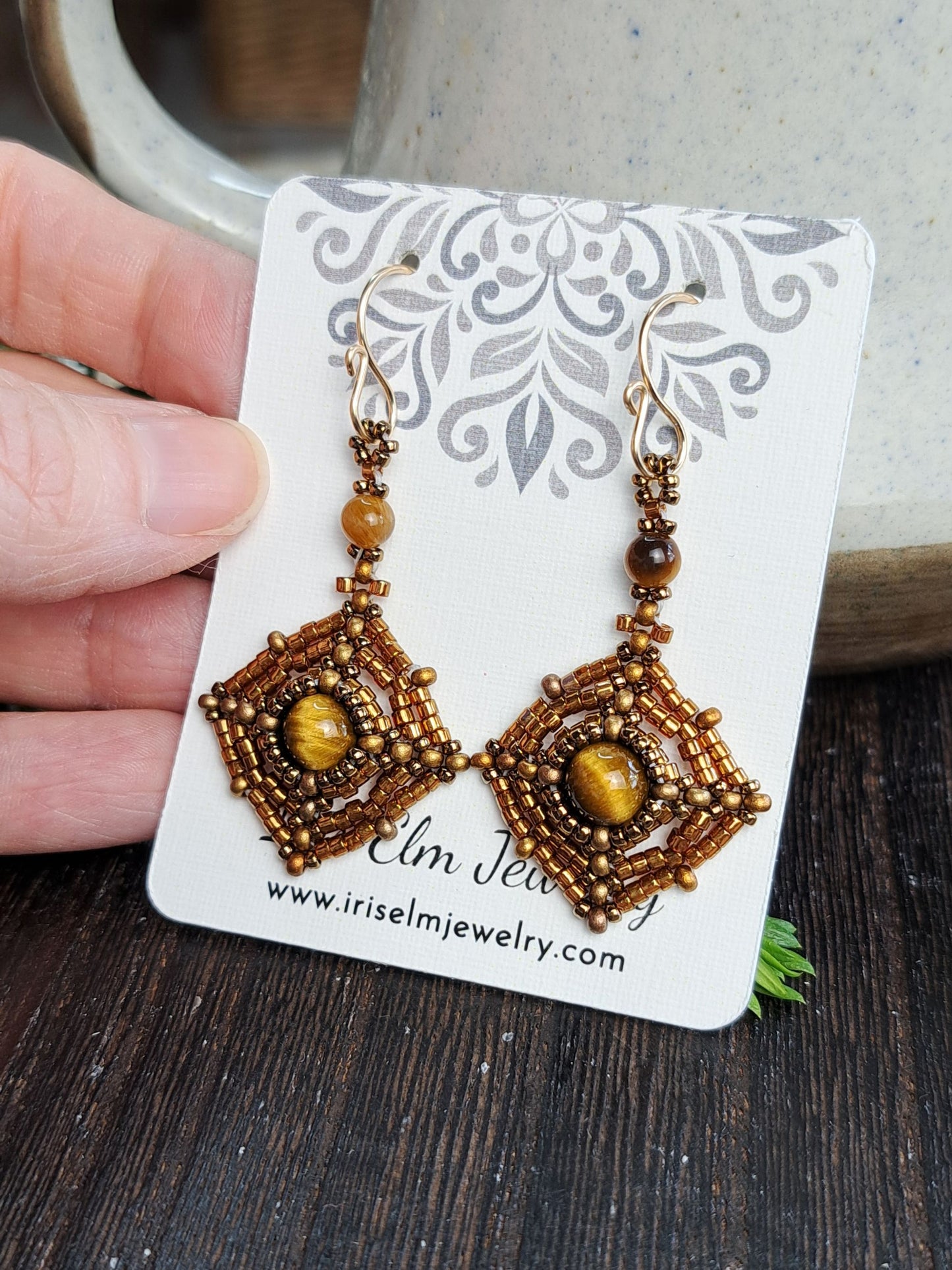 A pair of handmade beaded dangle earrings held on a white display card by a hand. The earrings feature intricate gold and brown seed bead geometric patterns with round amber-brown beads and central oval yellow-gold stones.