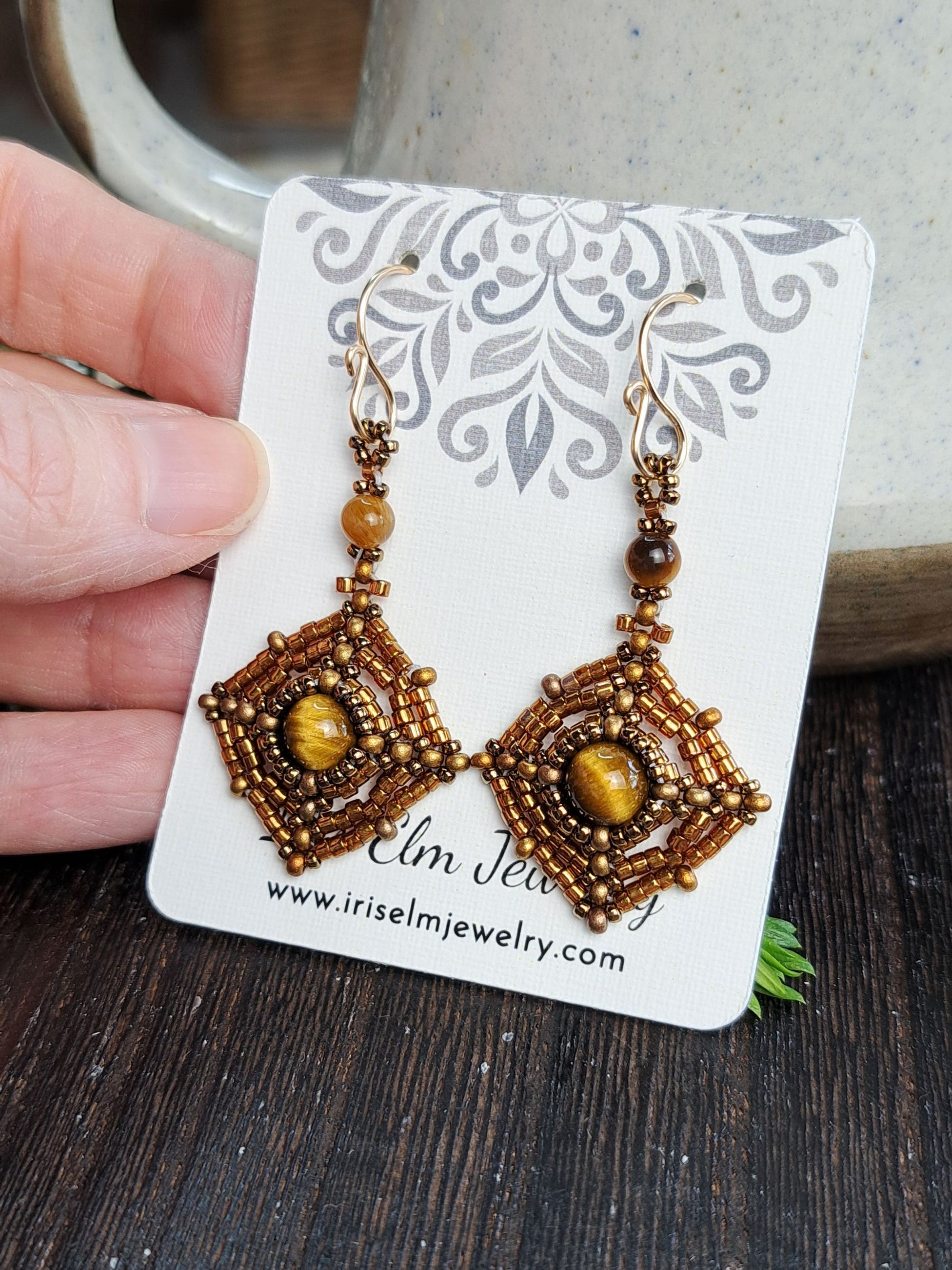 A pair of handmade beaded dangle earrings held on a white display card by a hand. The earrings feature intricate gold and brown seed bead geometric patterns with round amber-brown beads and central oval yellow-gold stones.
