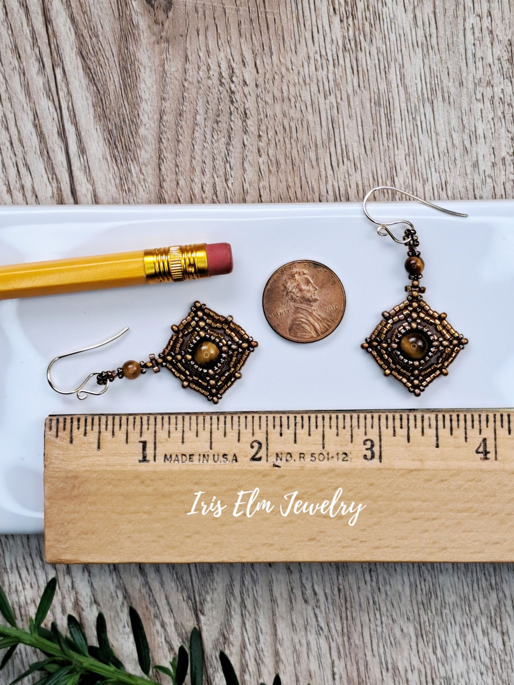 A pair of 2 inch long dangling geometric earrings with dark bronze and gold-toned metalwork, displayed on a white tray with a pencil, a penny, a wooden ruler, and greenery on a wood surface. The text 'Iris Elm Jewelry' appears on the ruler.