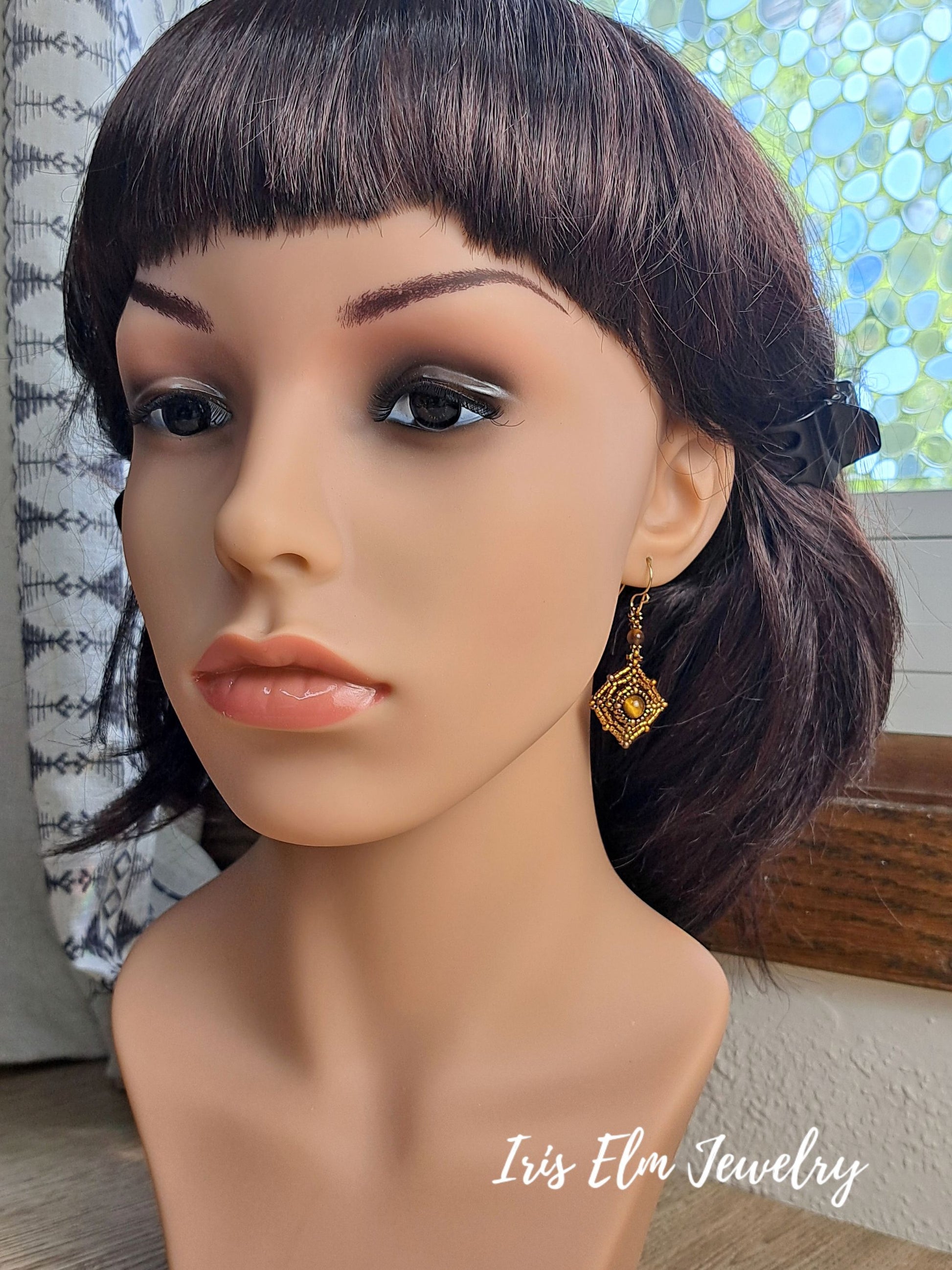 A gold geometric dangle earring with a central square-cut stone, displayed on a mannequin head with dark brown hair. The brand name 'Iris Elm Jewelry' is visible as a watermark in the bottom right corner.