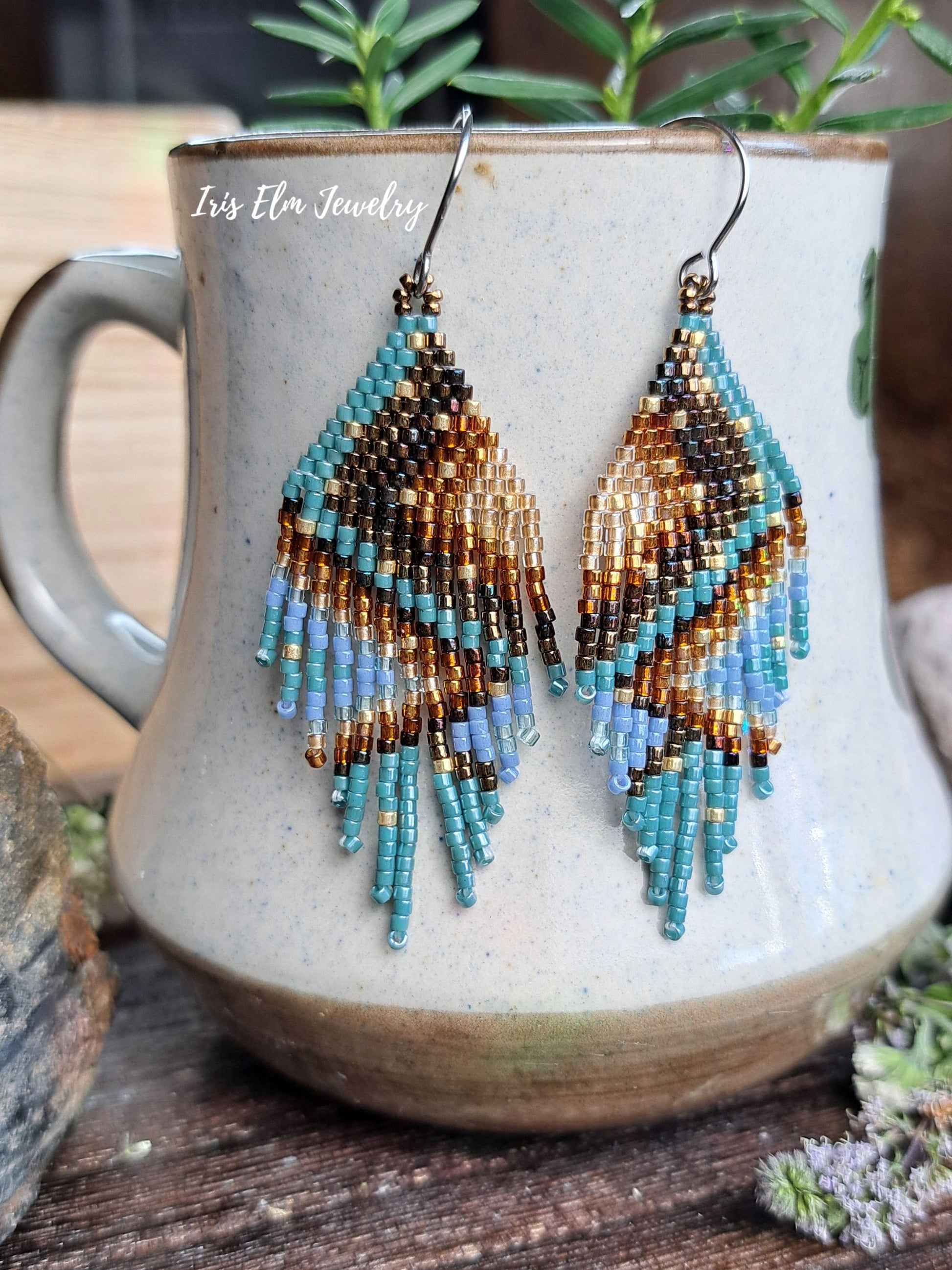 A pair of handmade beaded dangle earrings with a geometric pattern in turquoise, brown, black, and light blue, hanging from silver hooks in front of a ceramic mug. The mug has the text 'Iris Elm Jewelry' printed on it, which is listed as the visible brand.