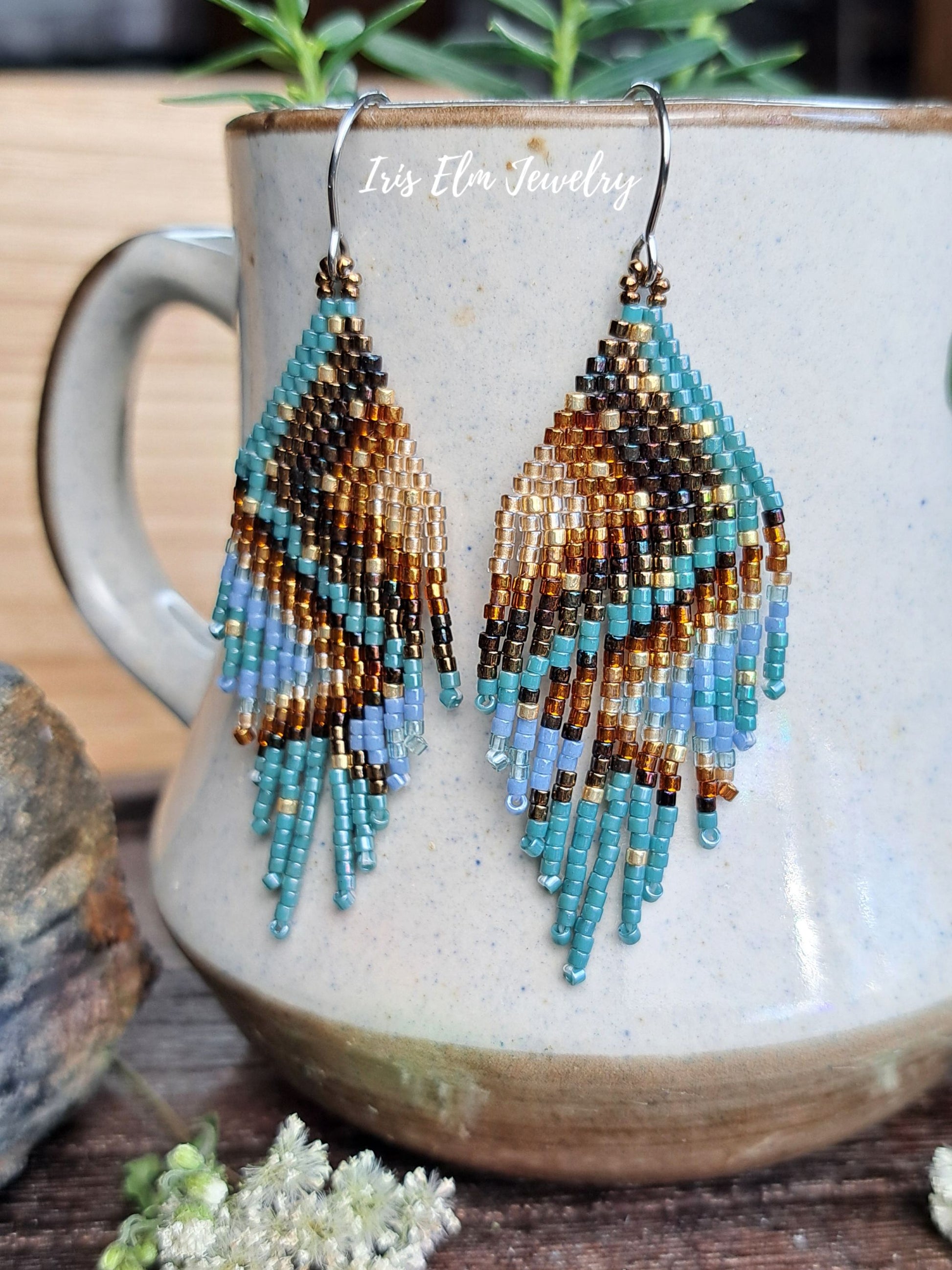 A pair of handmade beaded dangle earrings with a geometric pattern in brown, gold, turquoise, and light blue, hanging from silver hooks in front of a ceramic mug. The text 'Iris Elm Jewelry' is visible on the mug.