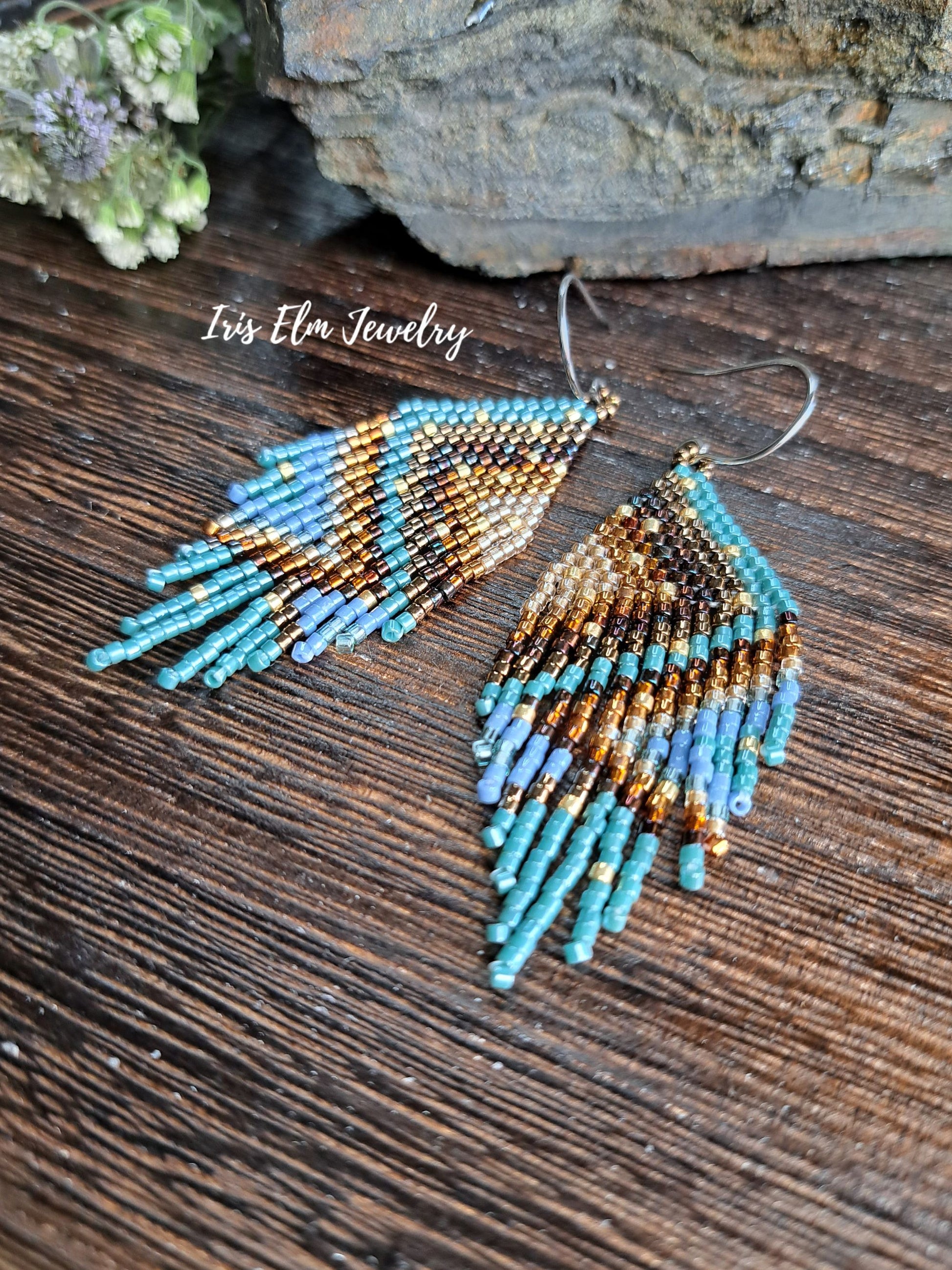 A pair of handmade beaded dangle earrings with a geometric pattern in turquoise, brown, and gold, resting on a dark wooden surface with a stone and greenery in the background. The text 'Iris Elm Jewelry' is visible on the image.