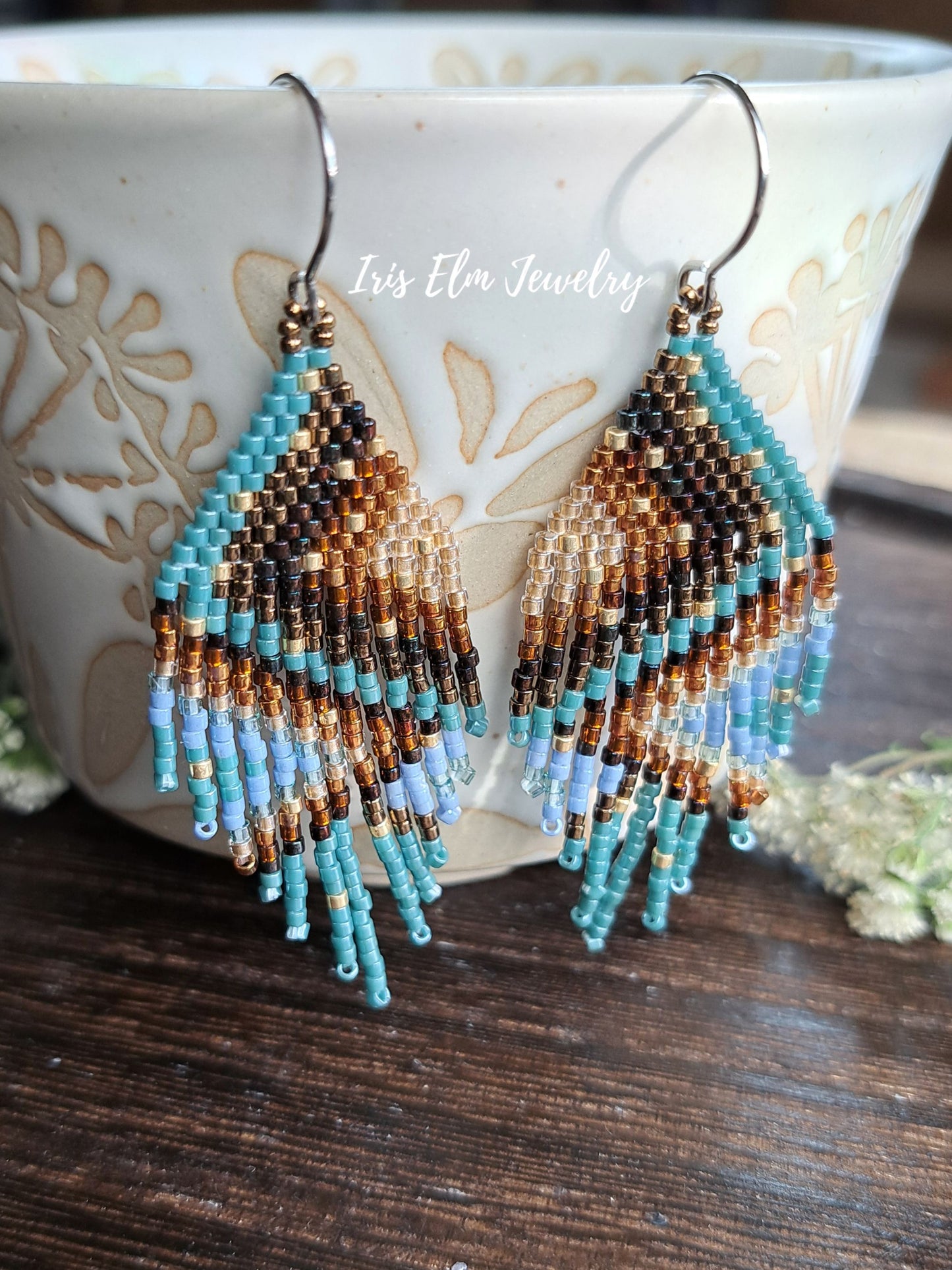 A pair of handmade beaded dangle earrings with a geometric pattern in turquoise, brown, black, and light blue, hanging in front of a ceramic cup on a dark wooden surface. The brand name 'Iris Elm Jewelry' is watermarked across the center.