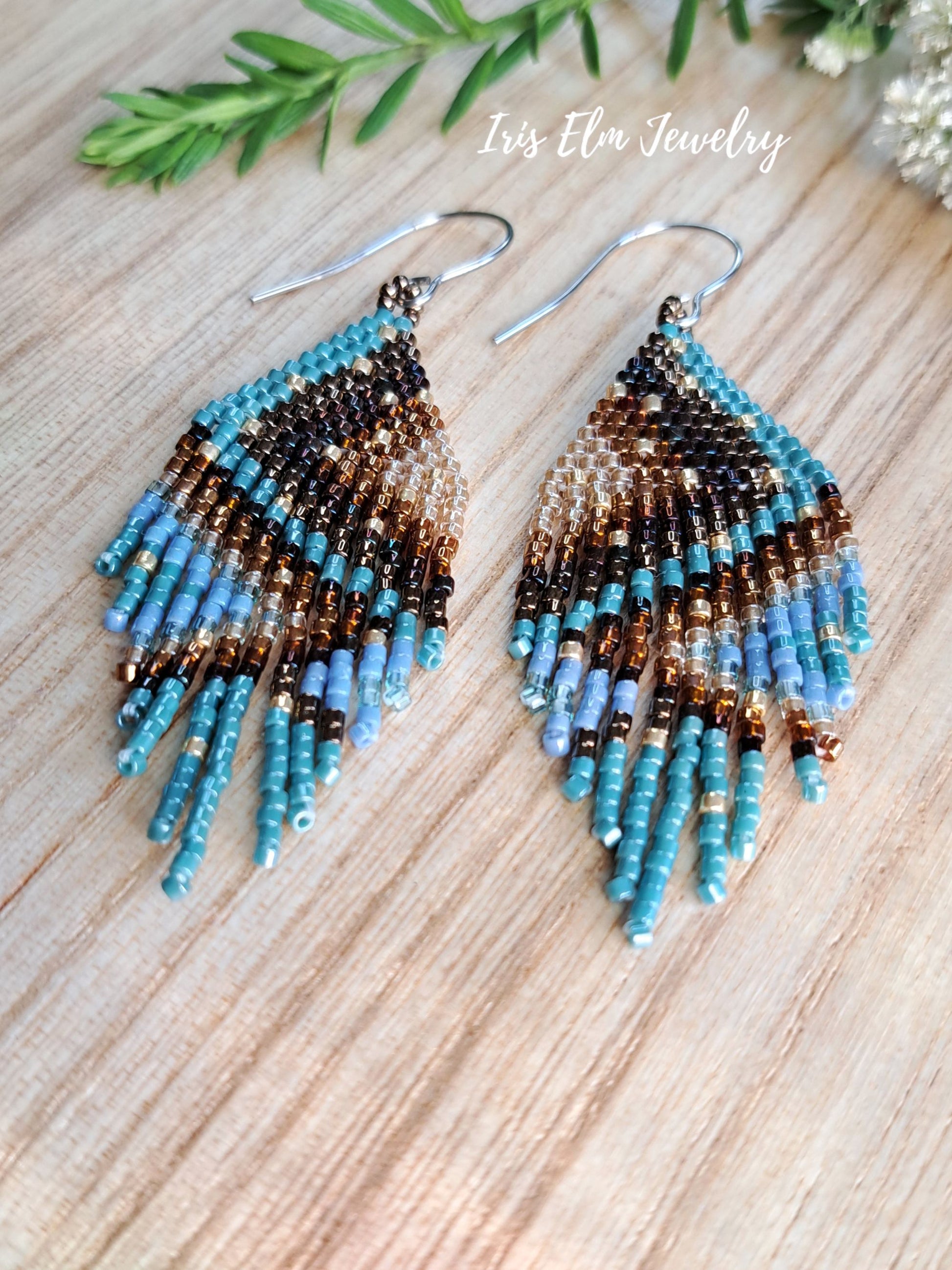 A pair of handmade beaded dangle earrings with a gradient design transitioning from dark brown and black at the top to light blue at the bottom, displayed on a light wood surface with green leaves in the corner. The text 'Iris Elm Jewelry' is visible at the top.