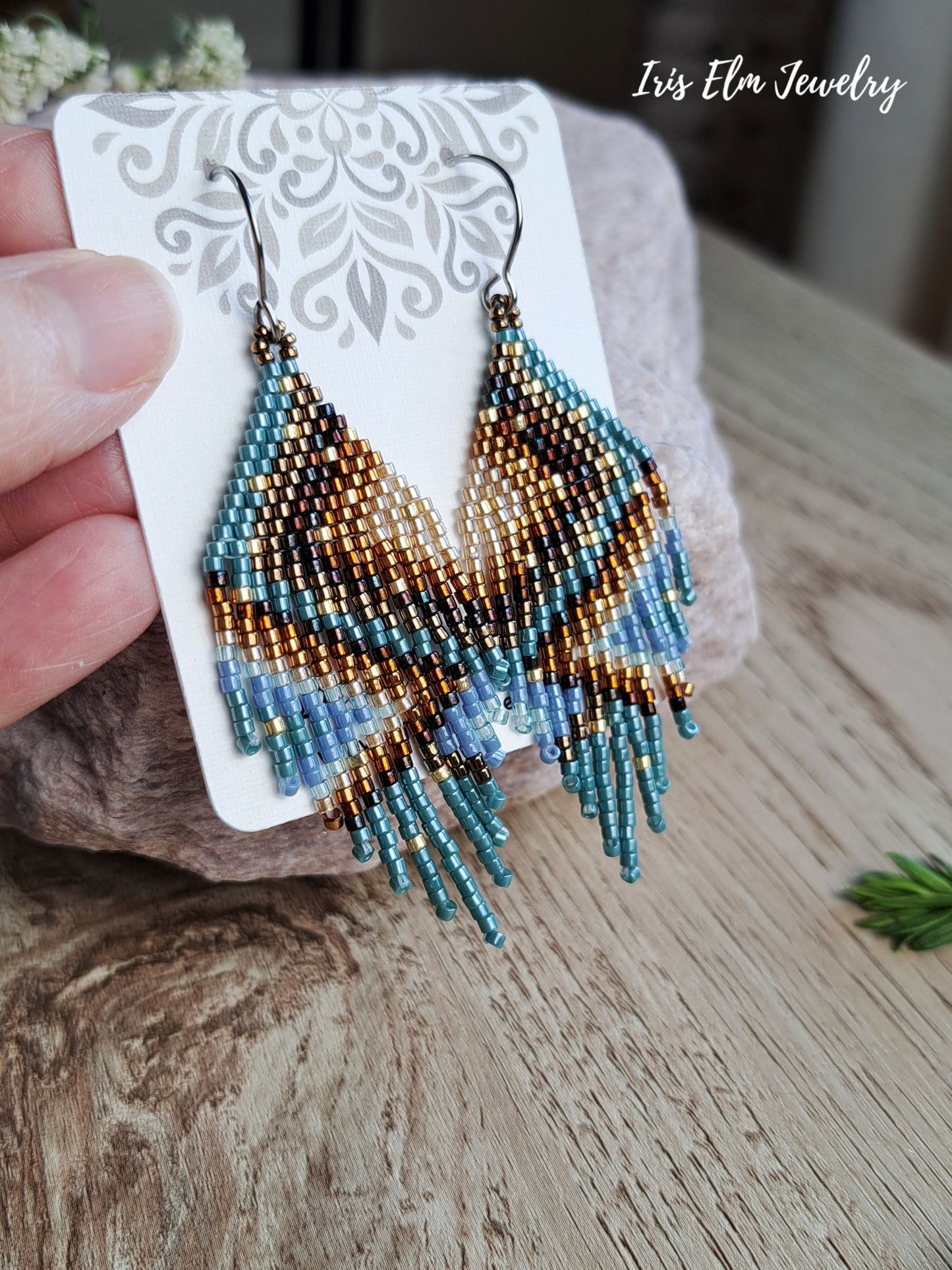 Hand-held beaded dangle earrings on a white display card, featuring a geometric pattern in turquoise, brown, and gold with silver hooks, set against a rustic wooden background.