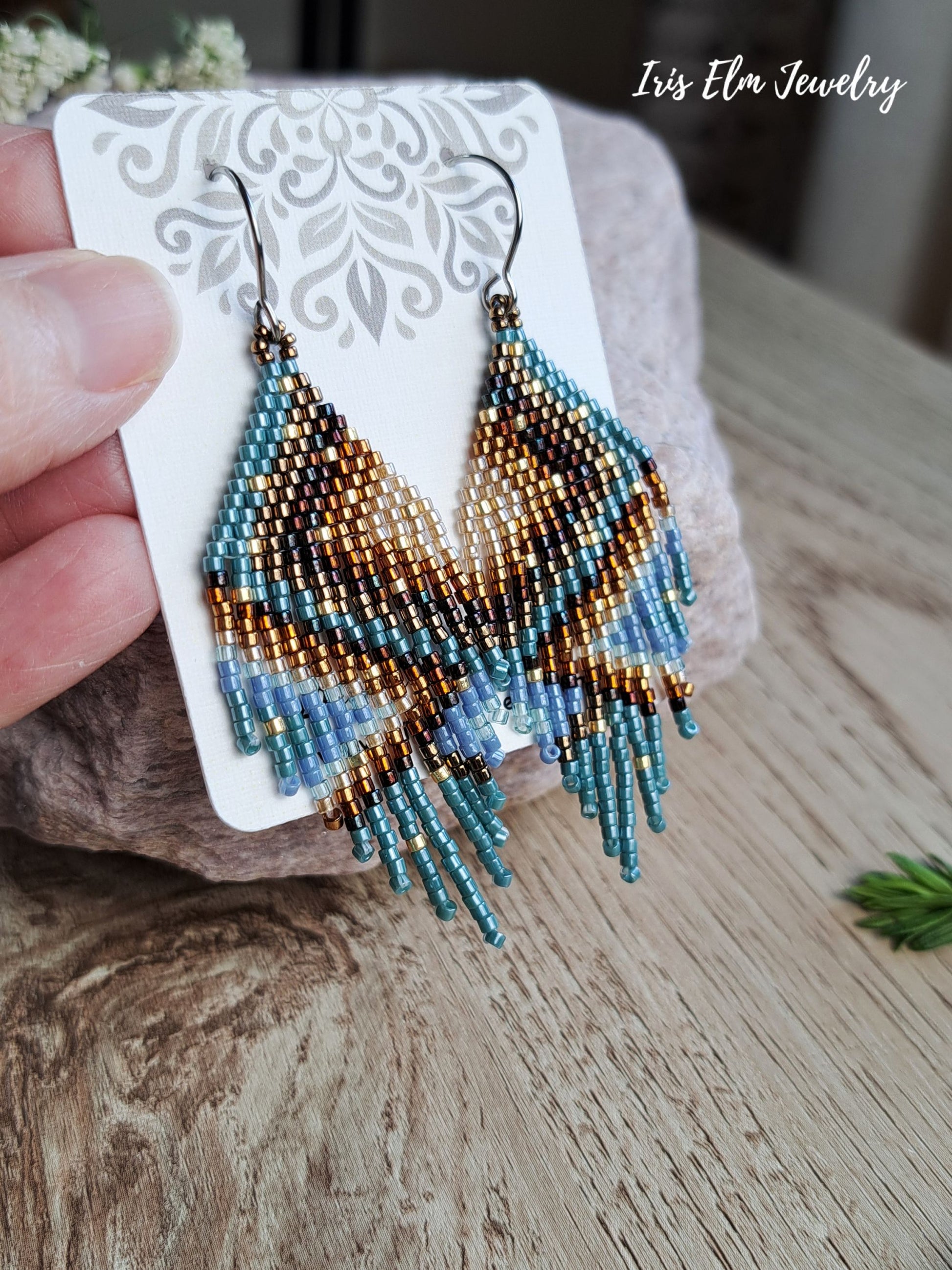 Hand-held beaded dangle earrings on a white display card, featuring a geometric pattern in turquoise, brown, and gold with silver hooks, set against a rustic wooden background.
