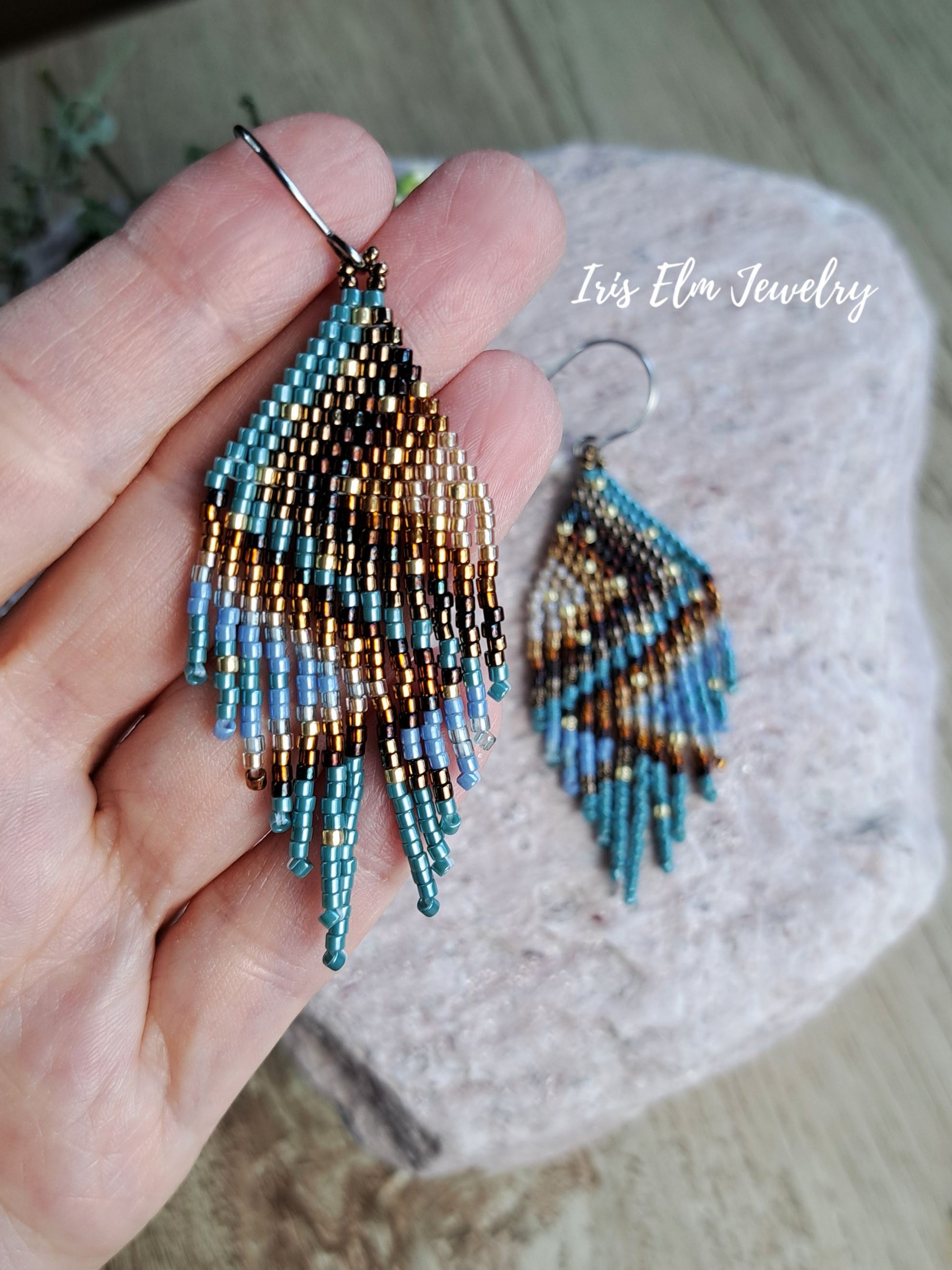 Hand holding a pair of beaded fringe earrings with a geometric pattern in turquoise, brown, and gold, with a matching earring resting on a stone background. The text 'Iris Elm Jewelry' is visible on the image.