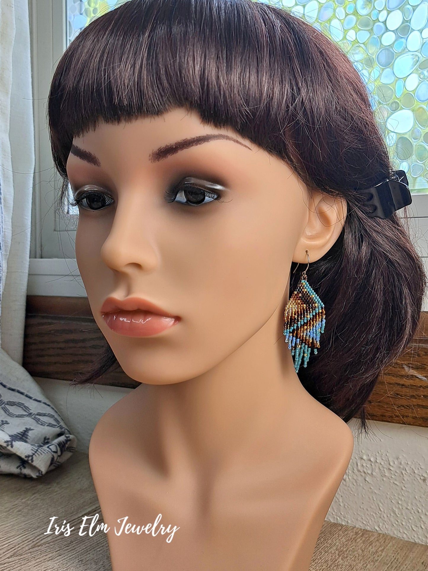A mannequin bust wearing a dark brown wig with straight bangs and a small black hair clip, shown indoors near a window. The mannequin is wearing a dangling beaded earring with blue, turquoise, orange, and brown tones.