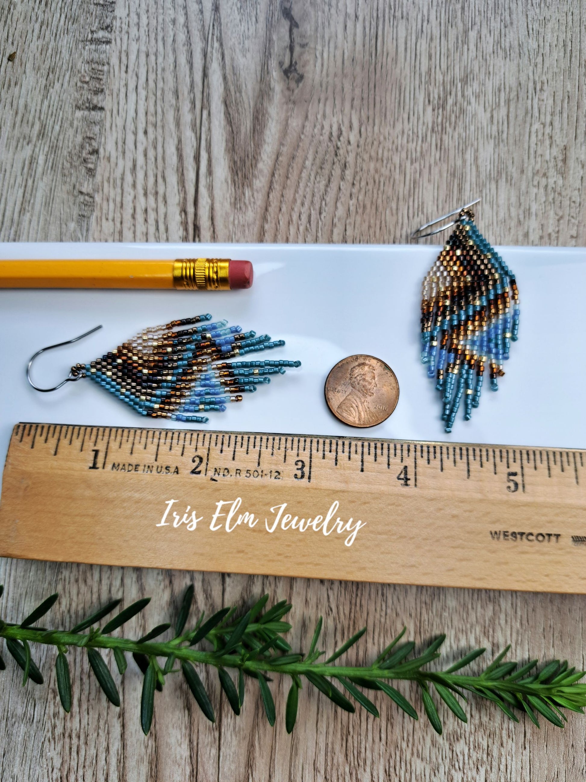 Two beaded 3 inch long dangle earrings displayed on a white tray with a pencil, penny, wooden ruler, and green sprig on a wood surface. The ruler is branded 'Iris Elm Jewelry'.