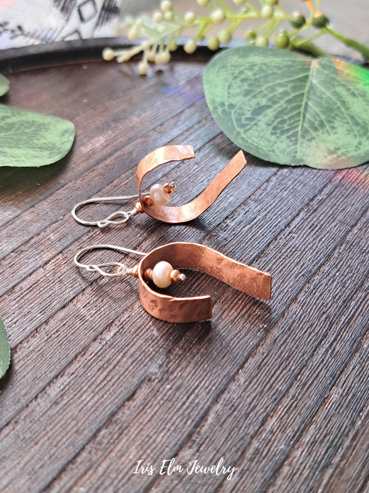 Copper and silver earrings with a pearl on a wooden surface with green leaves.