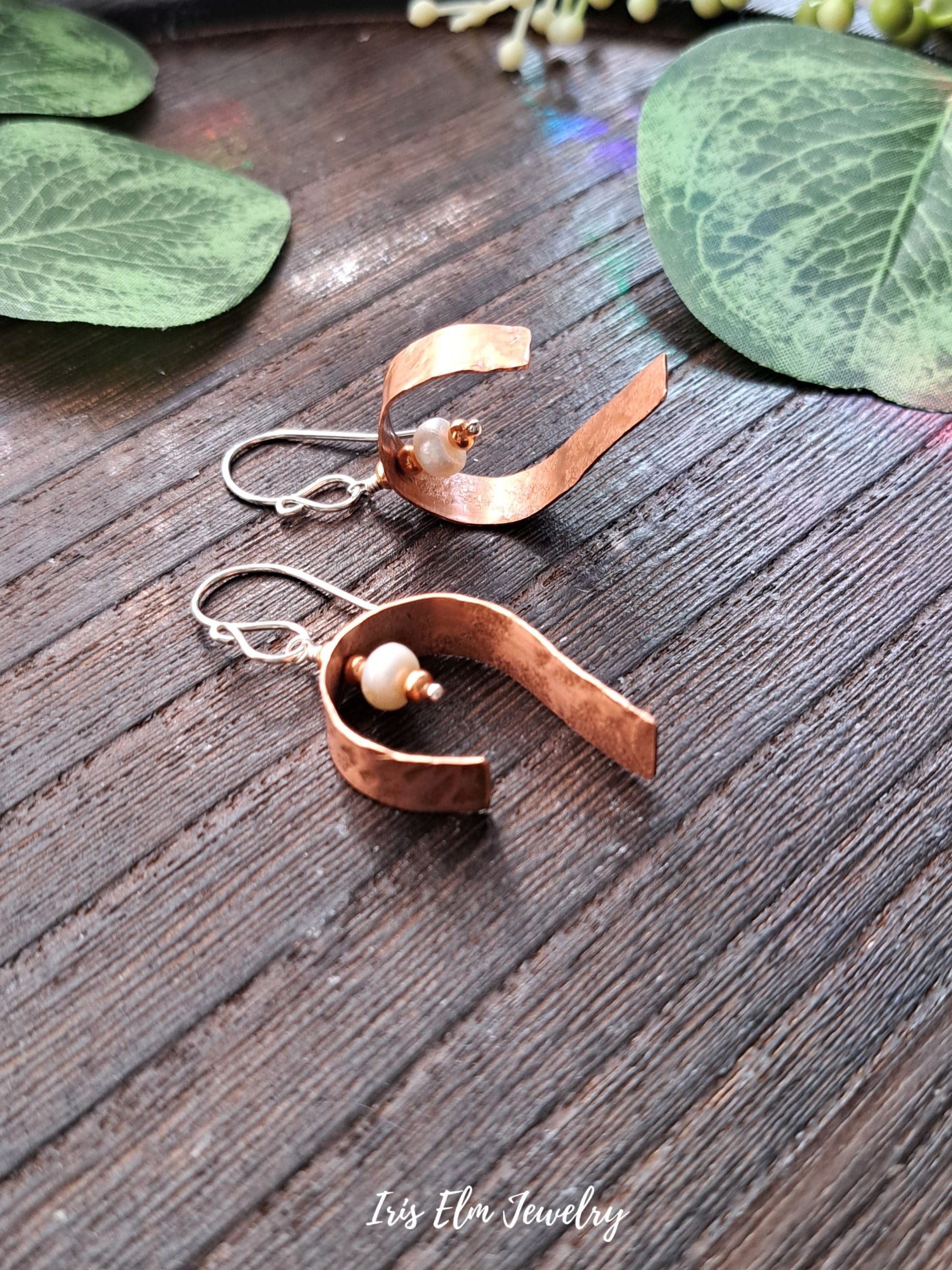 Copper earrings with a pearl on a wooden surface with green leaves.