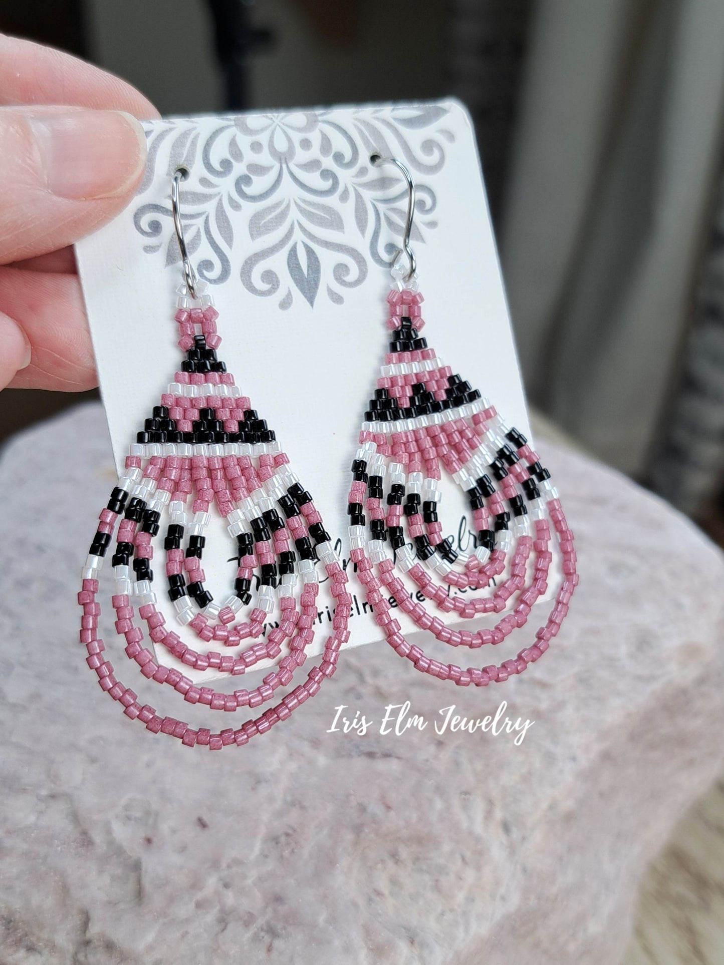 Pink, black, and white beaded dangle earrings on a white display card, held by a hand against a soft-focus gray background. The brand name 'Iris Elm Jewelry' is visible as a watermark.