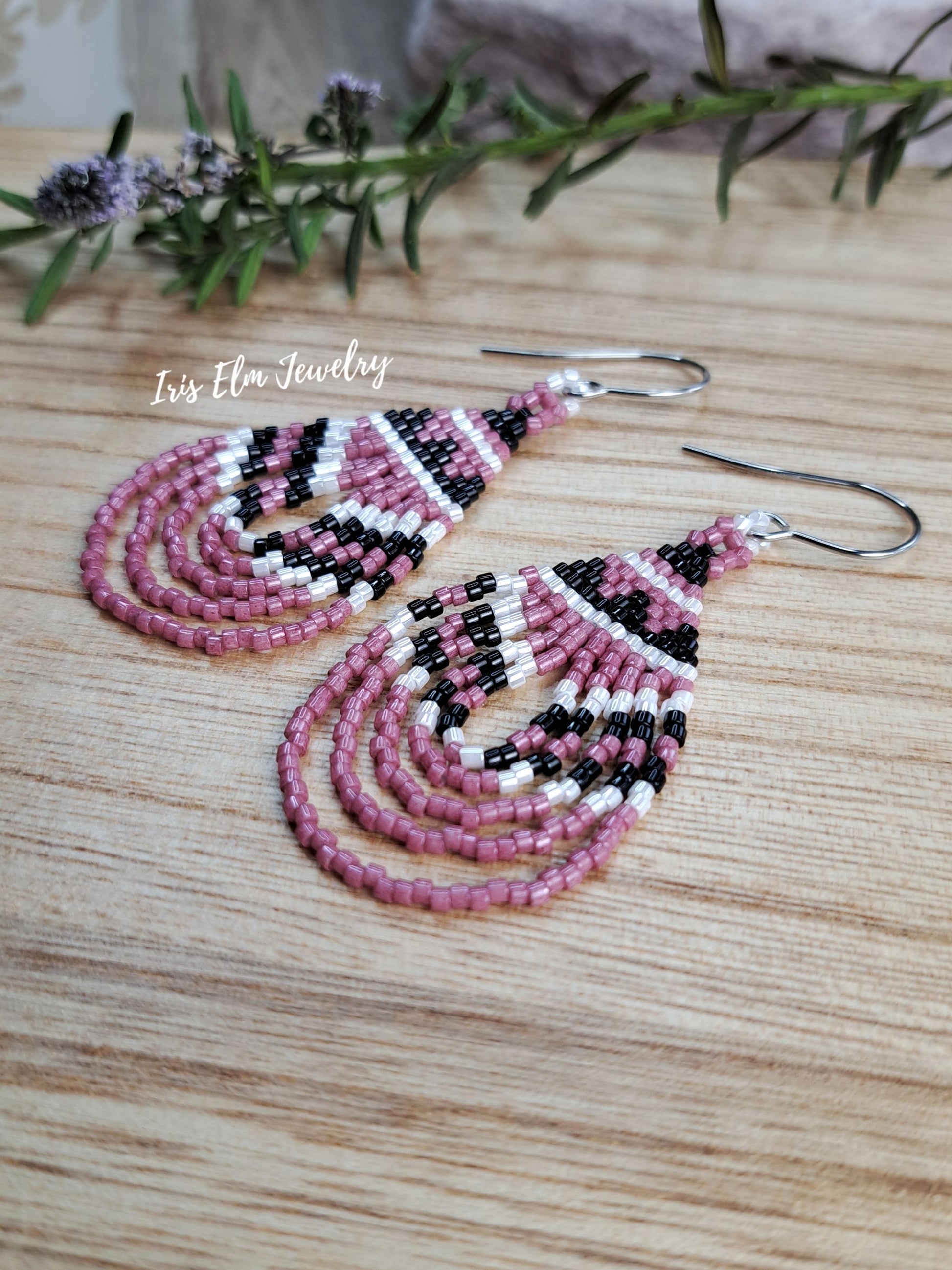 A pair of handmade beaded dangle earrings with a geometric pattern in pink, black, and white, hanging from silver fishhook wires on a light wood surface with a rosemary sprig in the background. The brand name 'Iris Elm Jewelry' is watermarked on the left.