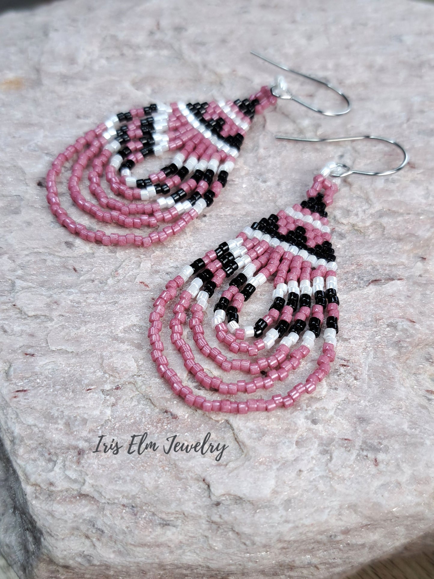 A pair of handmade beaded dangle earrings with pink, black, and white seed beads, displayed on a textured light gray stone surface. The brand name 'Iris Elm Jewelry' is printed on the stone.