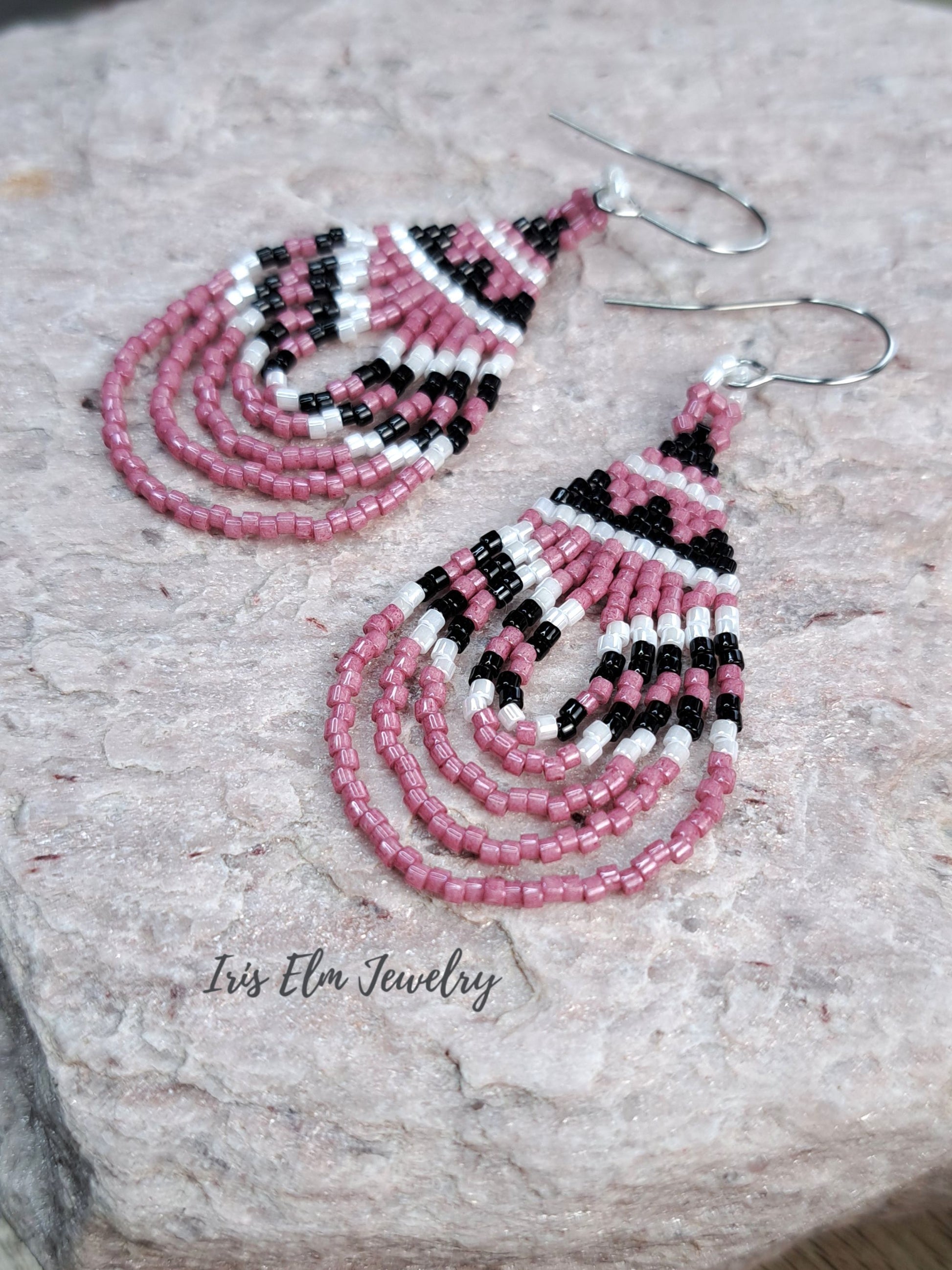 A pair of handmade beaded dangle earrings with pink, black, and white seed beads, displayed on a textured light gray stone surface. The brand name 'Iris Elm Jewelry' is printed on the stone.