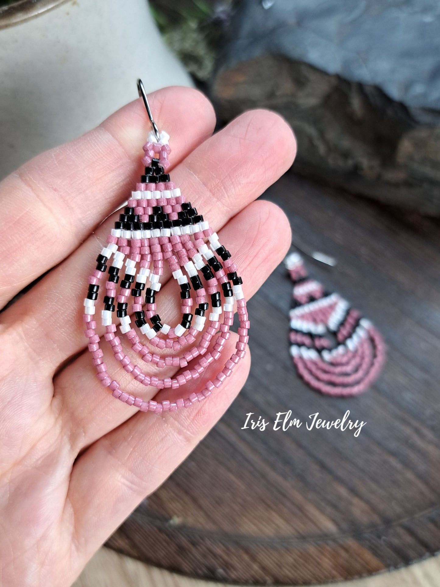 A hand holds a pink, black, and white beaded dangle earring, with a second matching earring resting on a dark wooden surface in the background. The brand name 'Iris Elm Jewelry' is watermarked on the bottom right.