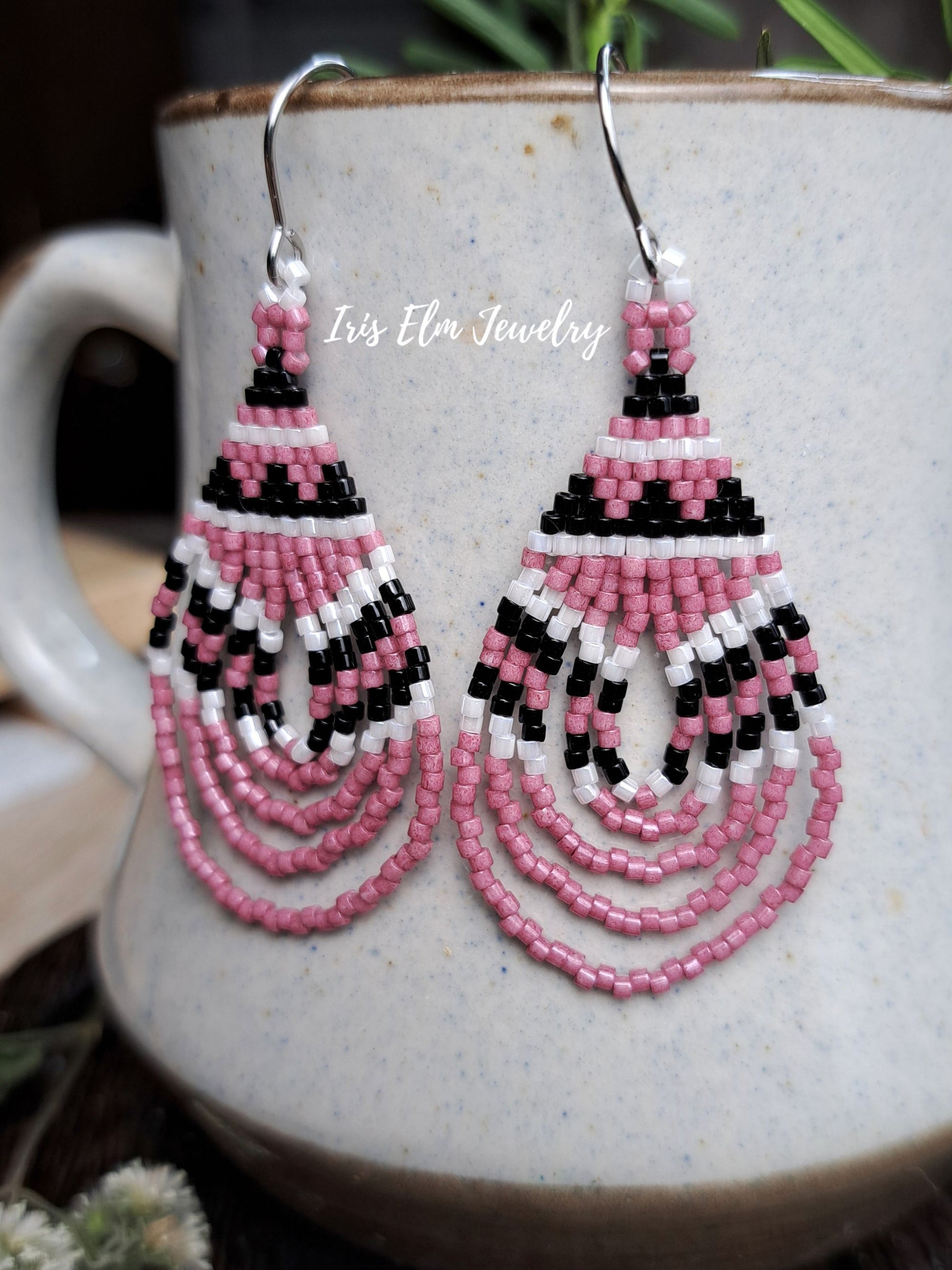 Pink, black, and white beaded dangle earrings with silver hooks, displayed on a speckled ceramic mug. The brand name 'Iris Elm Jewelry' is watermarked across the center.