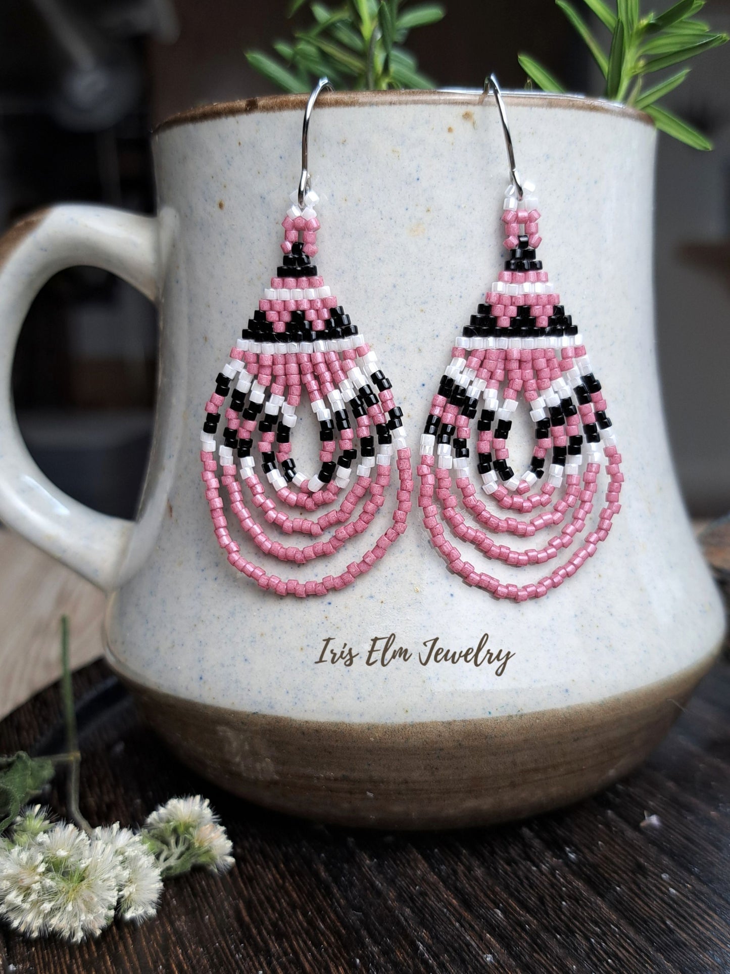 A pair of handmade beaded dangle earrings in pink, black, and white, hanging from silver hooks on a ceramic mug. The brand name 'Iris Elm Jewelry' is printed on the front of the mug.