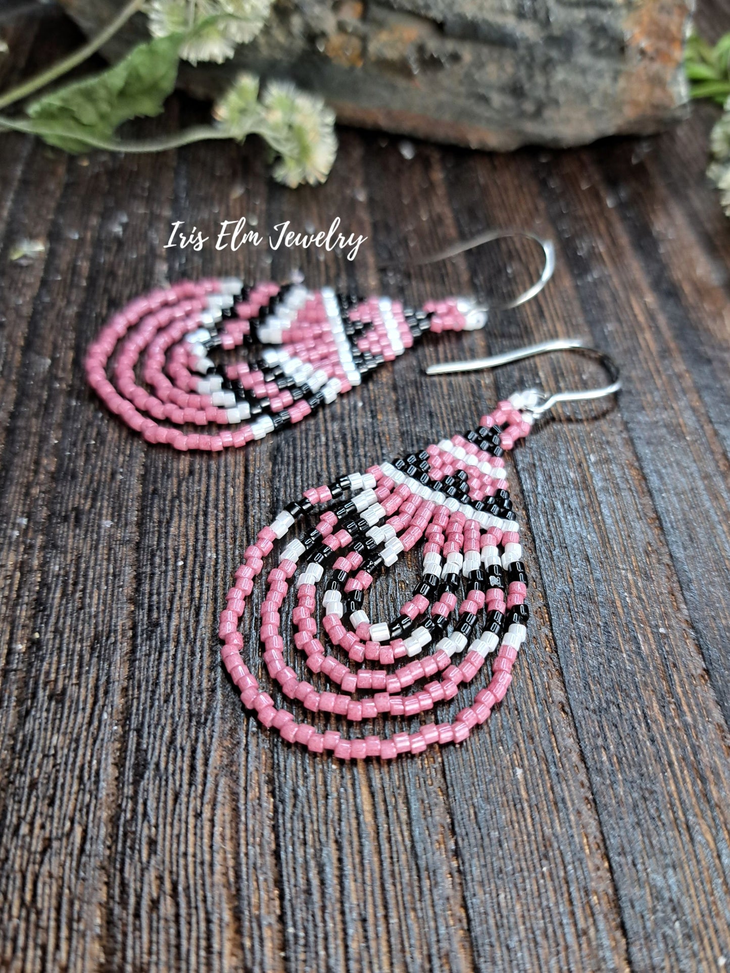 Pink, black, and white beaded teardrop dangle earrings with silver fishhook wires, displayed on a dark rustic wooden surface with a brand watermark 'Iris Elm Jewelry'.