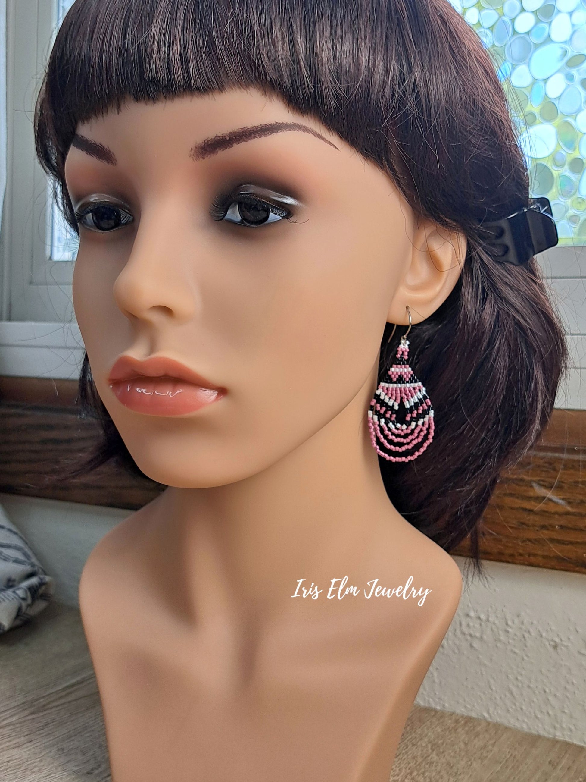 A close-up of a mannequin bust wearing a dangling beaded earring. The earring features layered rows of pink, white, and dark beads in a fan or tassel-like design.