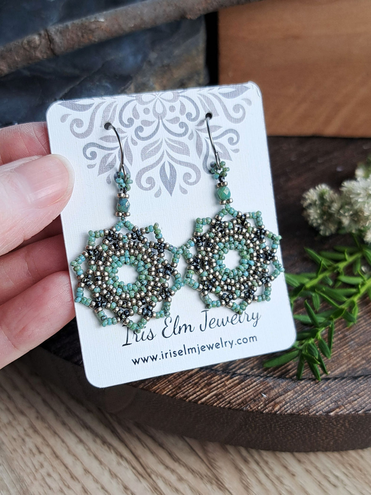 A hand holds a white jewelry display card featuring a pair of intricate beaded earrings. The earrings have a silver-toned geometric starburst design with turquoise and dark gray beads, hanging from silver hooks.