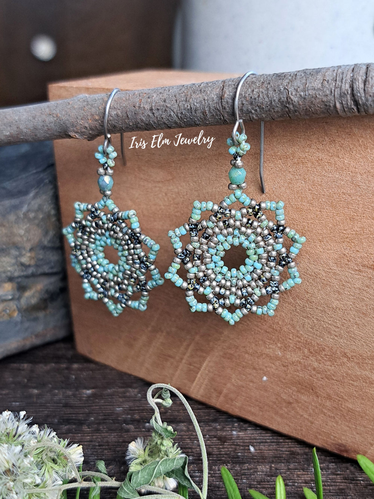 Pair of handmade beaded dangle earrings with turquoise, silver, and black seed beads, hanging from a rustic branch over a wooden block. The brand name 'Iris Elm Jewelry' is watermarked across the center.