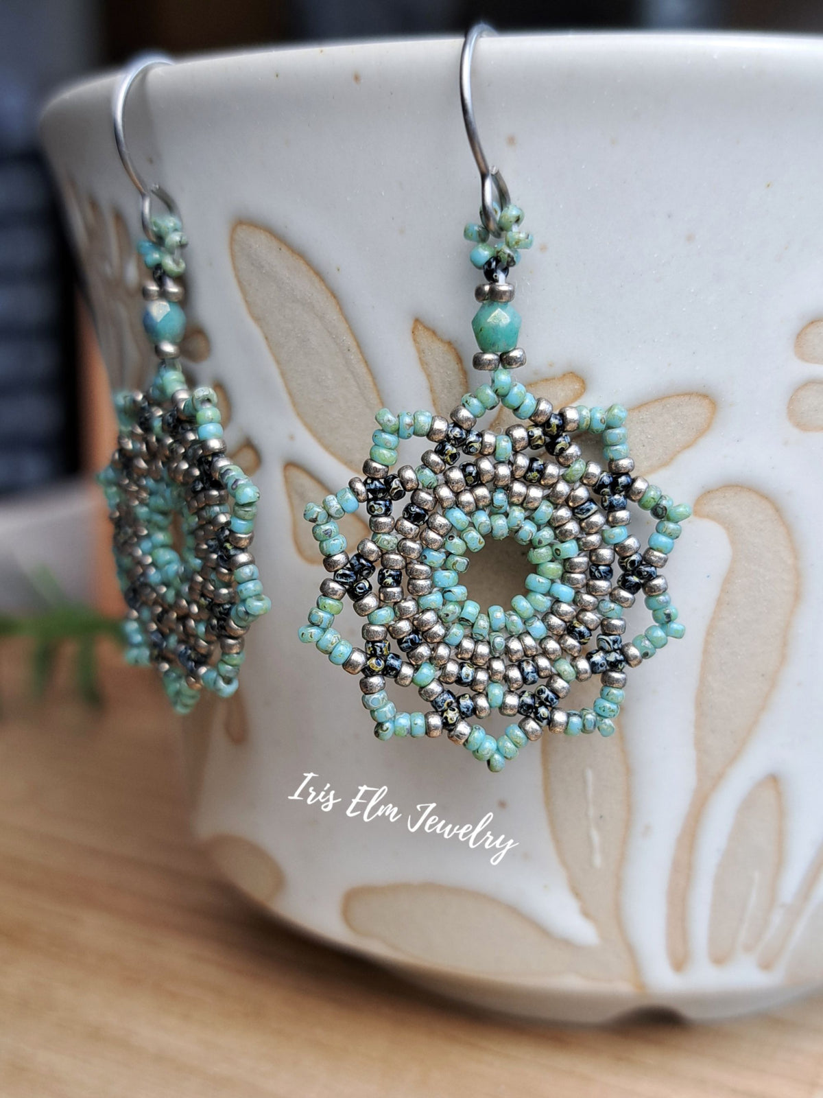 A pair of handmade beaded dangle earrings with intricate silver, black, and turquoise beadwork, hanging in front of a white ceramic mug. The brand name 'Iris Elm Jewelry' is watermarked on the mug.
