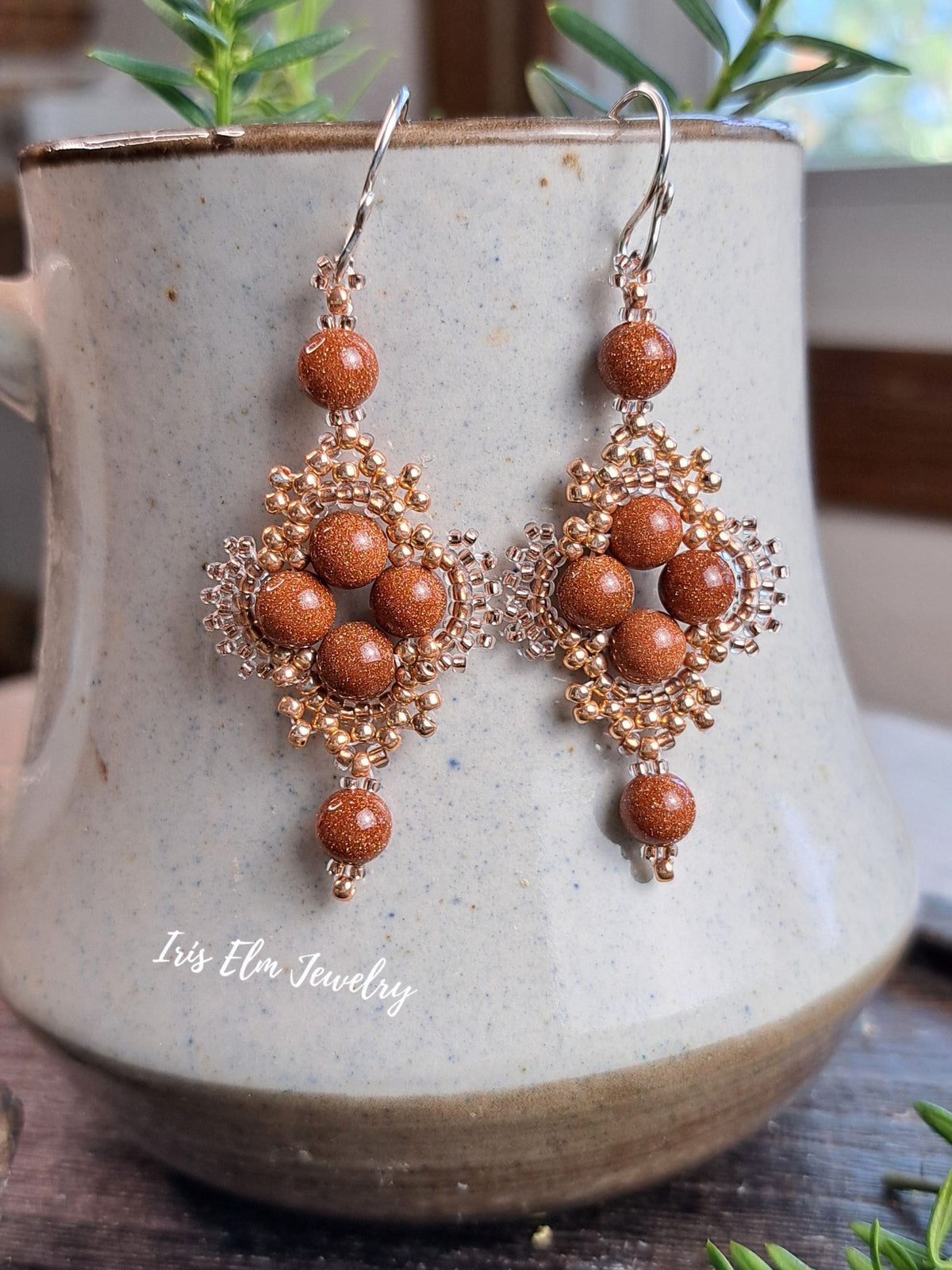 A pair of bohemian-style dangle earrings with copper-toned filigree and round brown goldstone beads, displayed on a speckled ceramic mug with a watermark reading 'Iris Elm Jewelry'.