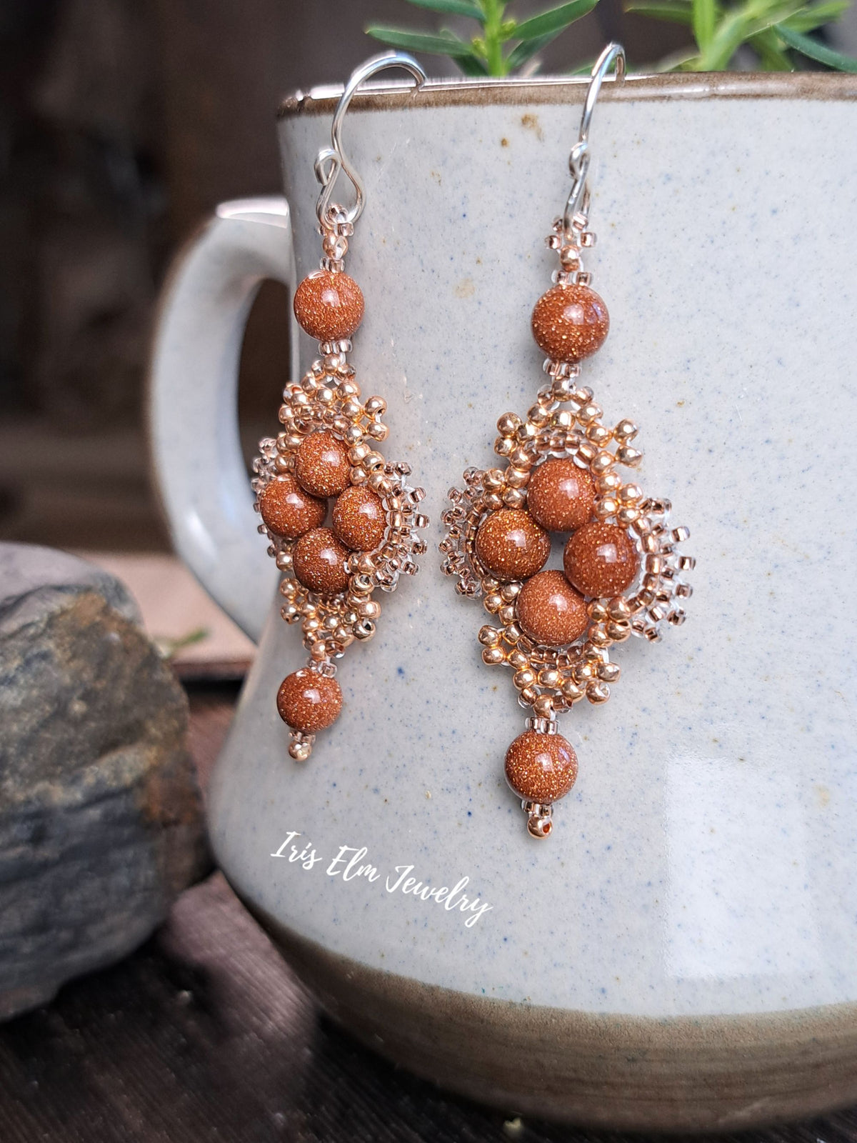 A pair of dangling earrings with copper-toned beaded filigree and round brown goldstone beads, hanging from silver fishhook wires in front of a speckled ceramic mug. The brand name 'Iris Elm Jewelry' is printed on the mug.