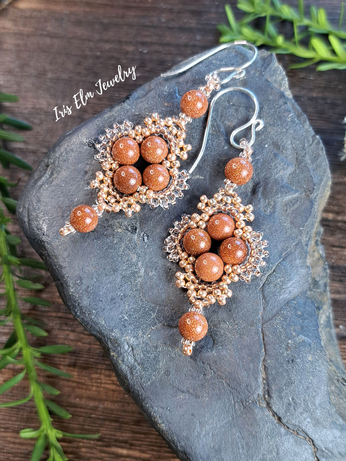 A pair of handmade dangle earrings with copper-toned beads, clear crystal filigree, and silver fishhook wires, displayed on a dark gray slate stone with green pine branches on a wooden surface. The brand name 'Iris Elm Jewelry' is watermarked on the top left.
