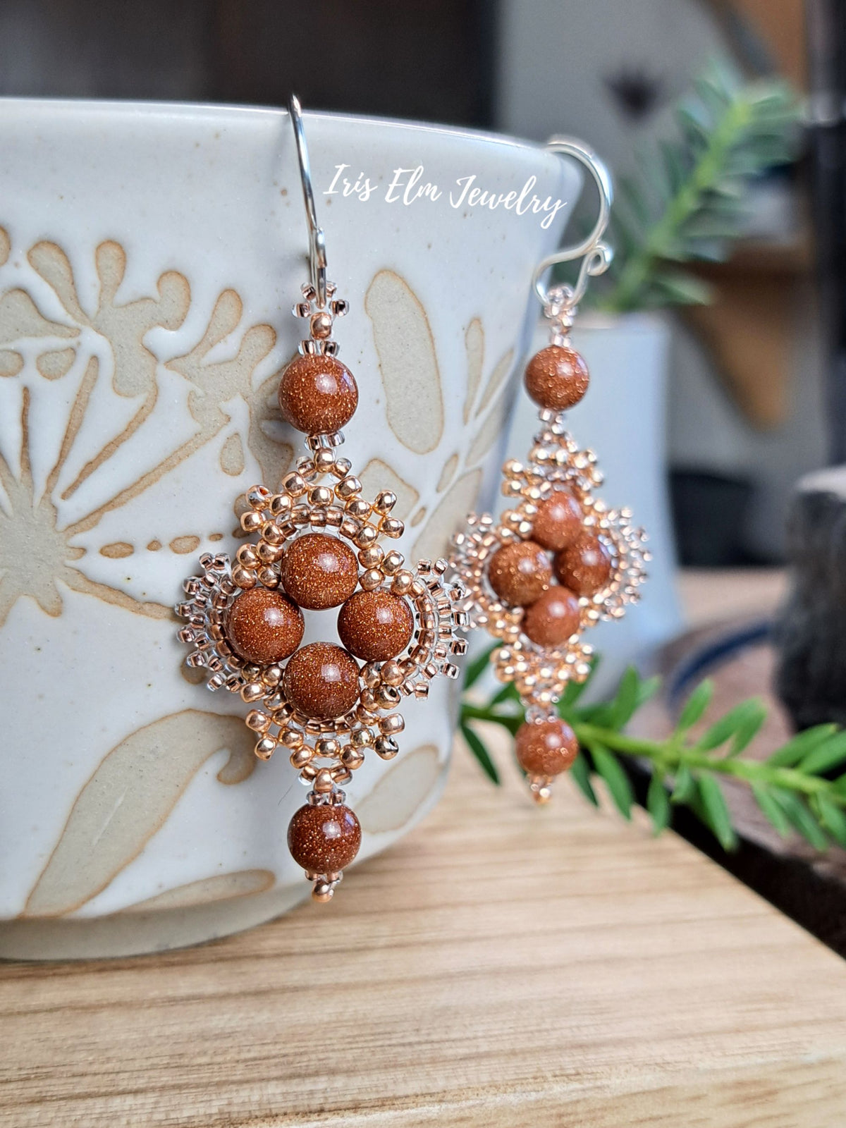 A pair of dangling earrings with coppery brown stone beads and rose gold-toned metalwork, displayed in front of a ceramic mug on a wooden surface with a blurred indoor background.